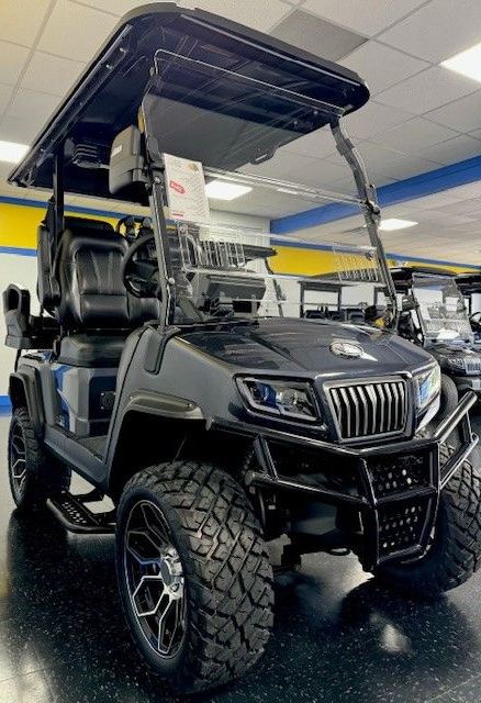 Black, lifted golf cart with large tires, a roof, and a front brush guard is displayed in a showroom.