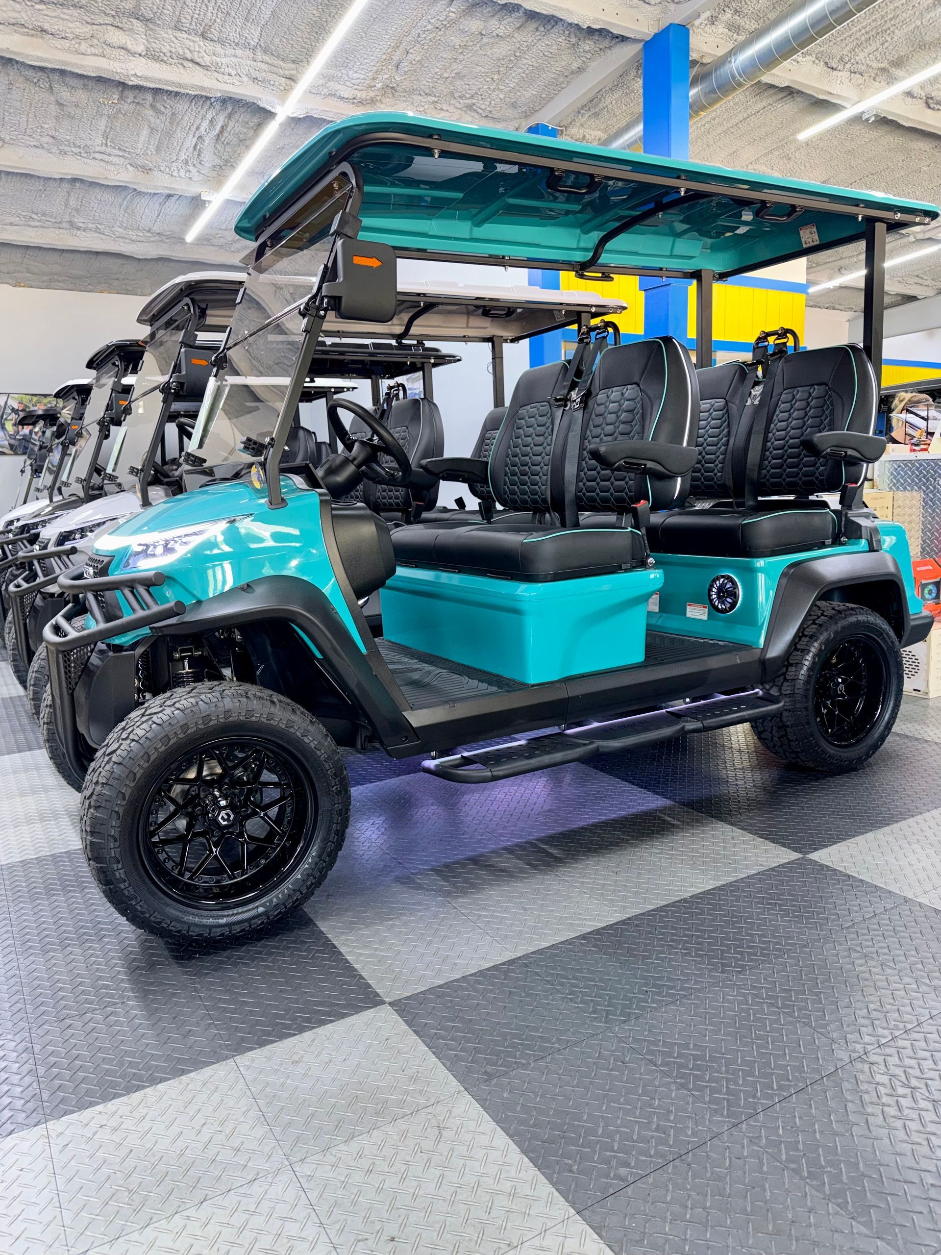 Turquoise golf cart with black seats and roof, parked indoors on a checkered showroom floor.