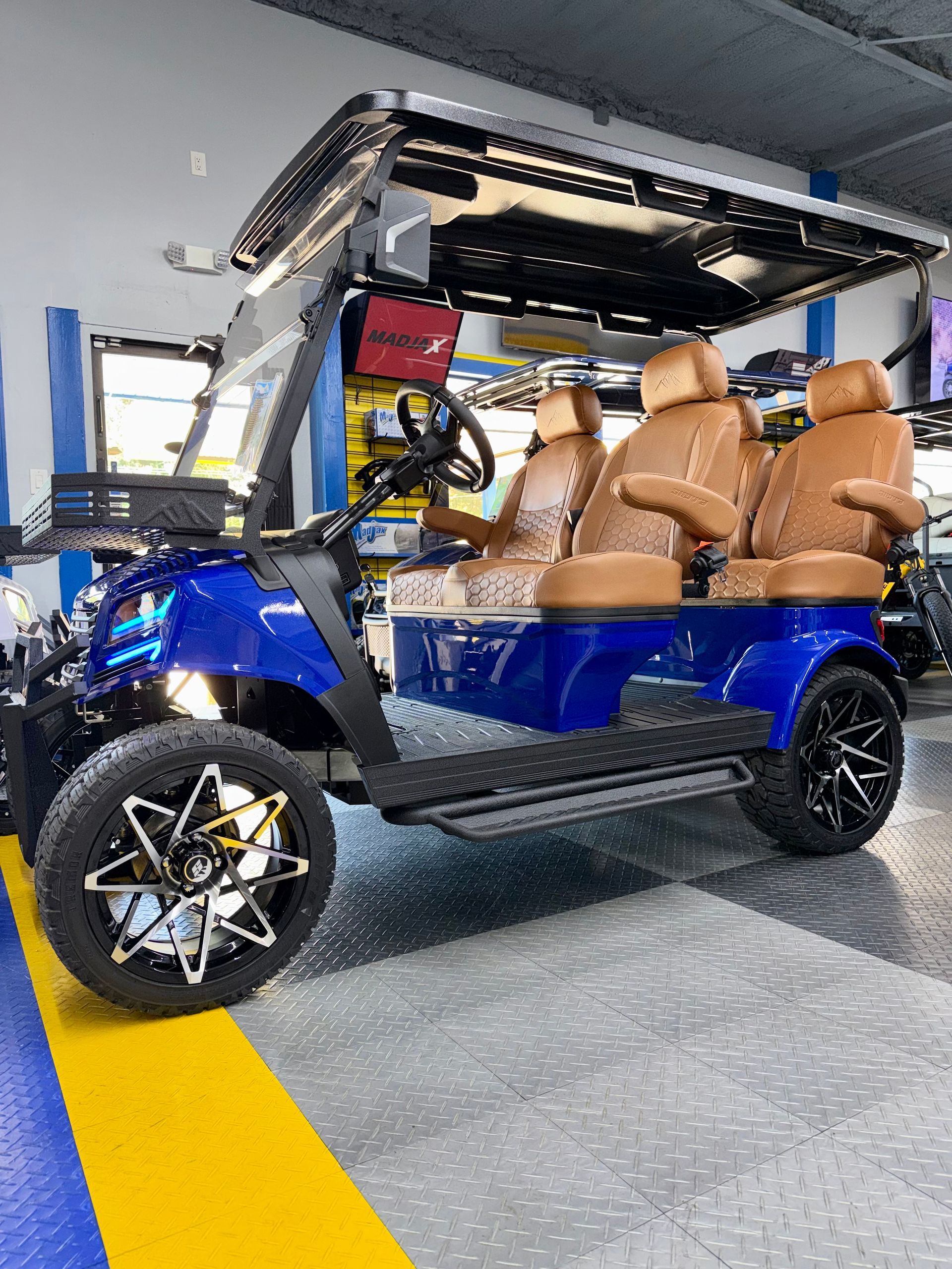 Blue off-road golf cart with tan seats parked indoors, lifted front hood open and showing custom wheels.