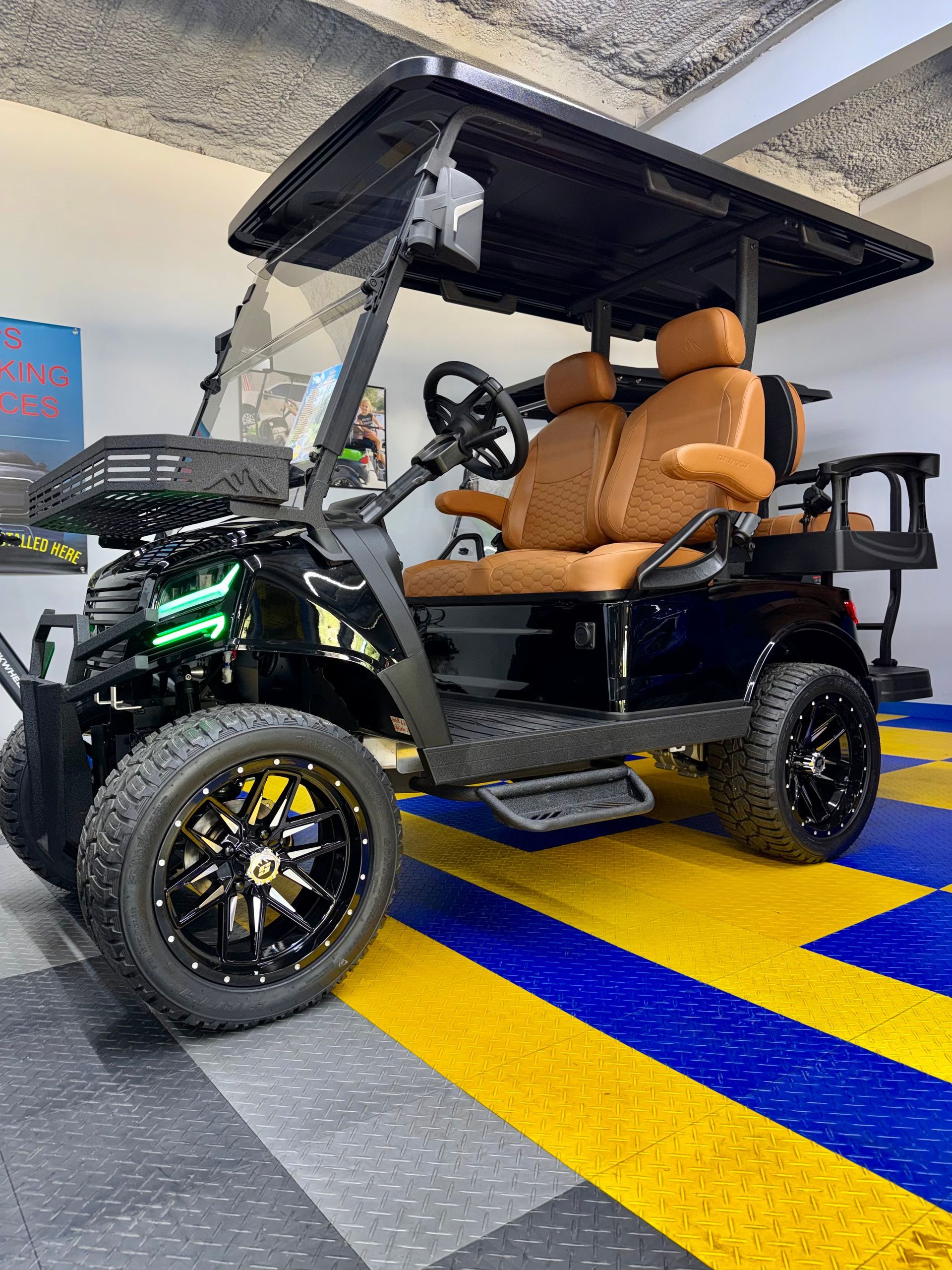 Black electric utility cart with tan seats and lifted wheels parked indoors on a gray floor with blue and yellow stripes