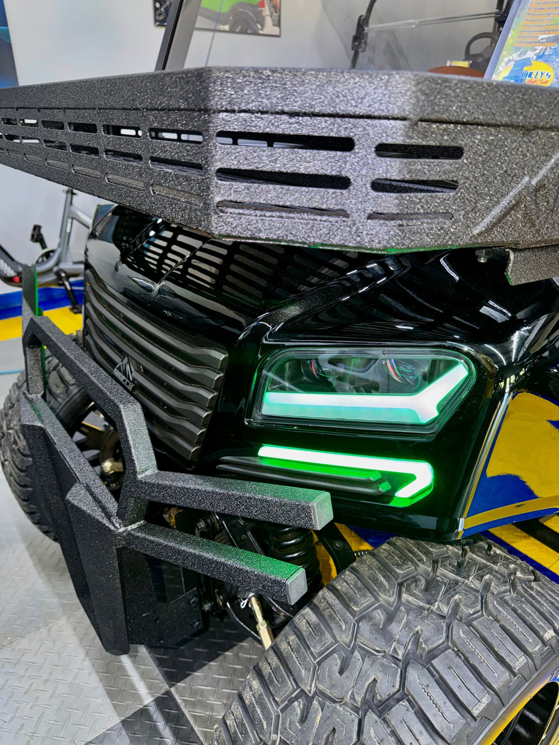 Modified black off-road vehicle front with green LED lights and chunky tire, indoors on a lift