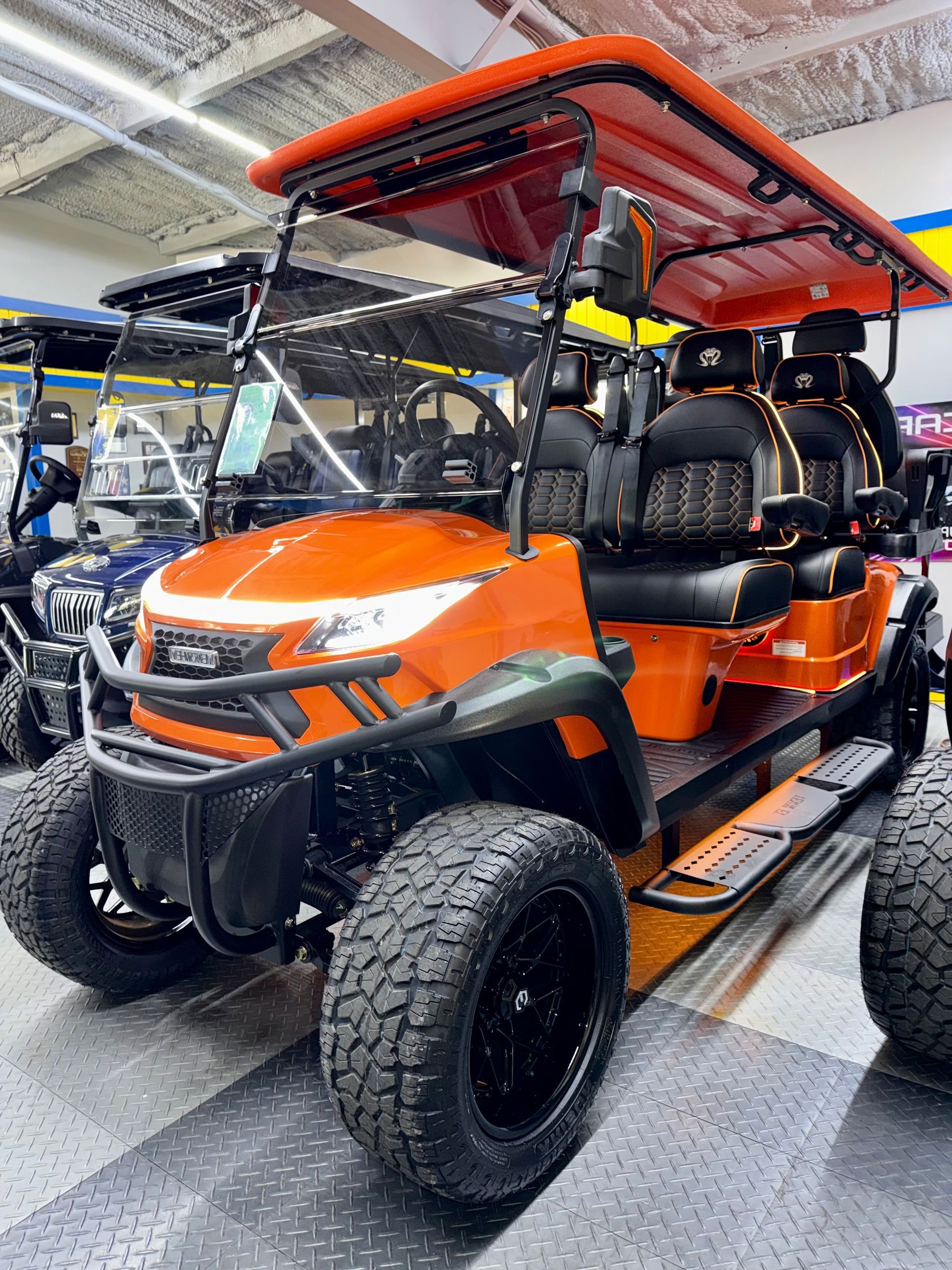 Orange off-road utility vehicle with black roll cage, parked indoors on a showroom floor.