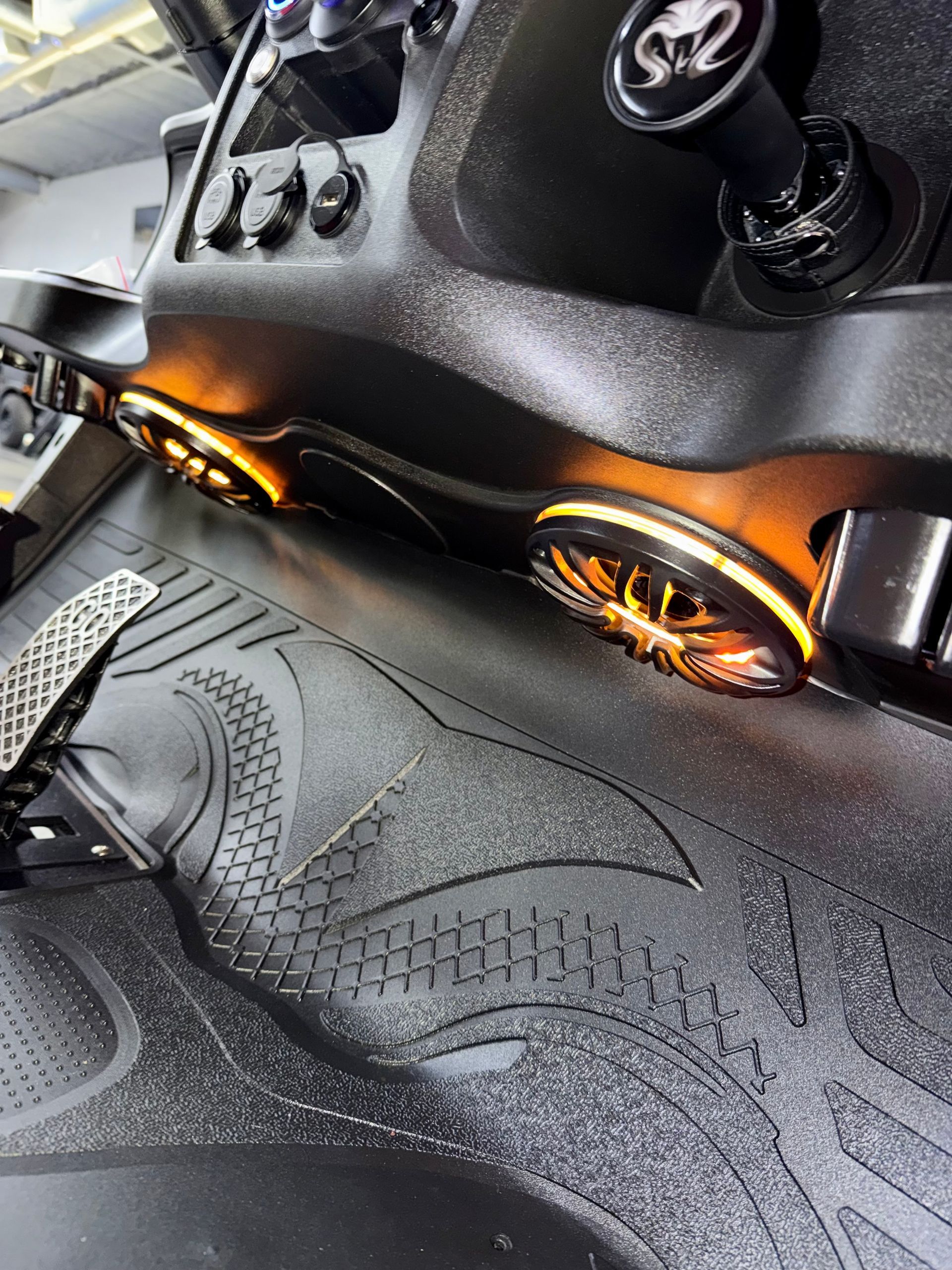 Black car dashboard with glowing orange gauges and manual gear shifter over a patterned floor mat