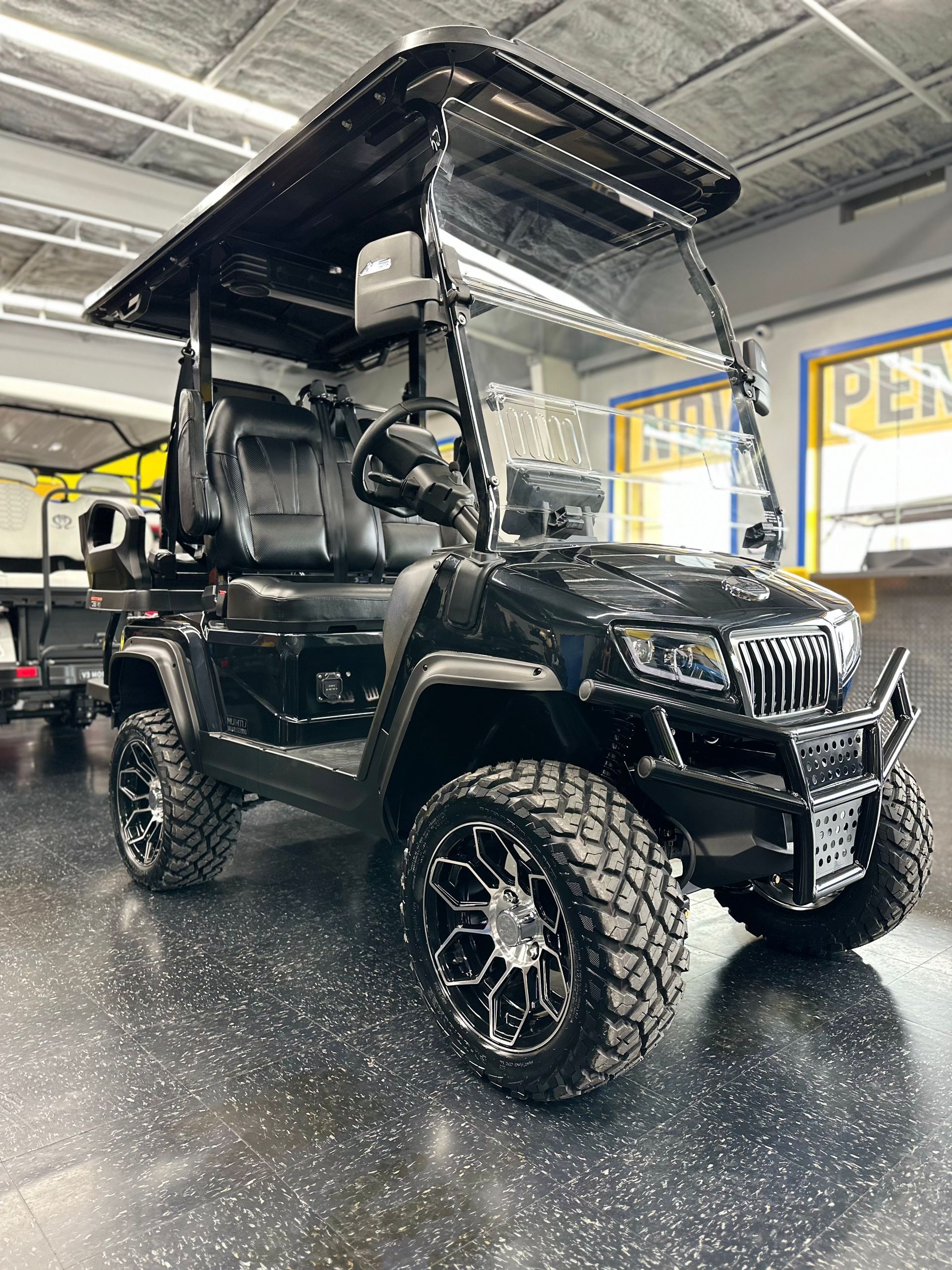 Black golf cart with large wheels and a roof, parked indoors on a reflective surface.