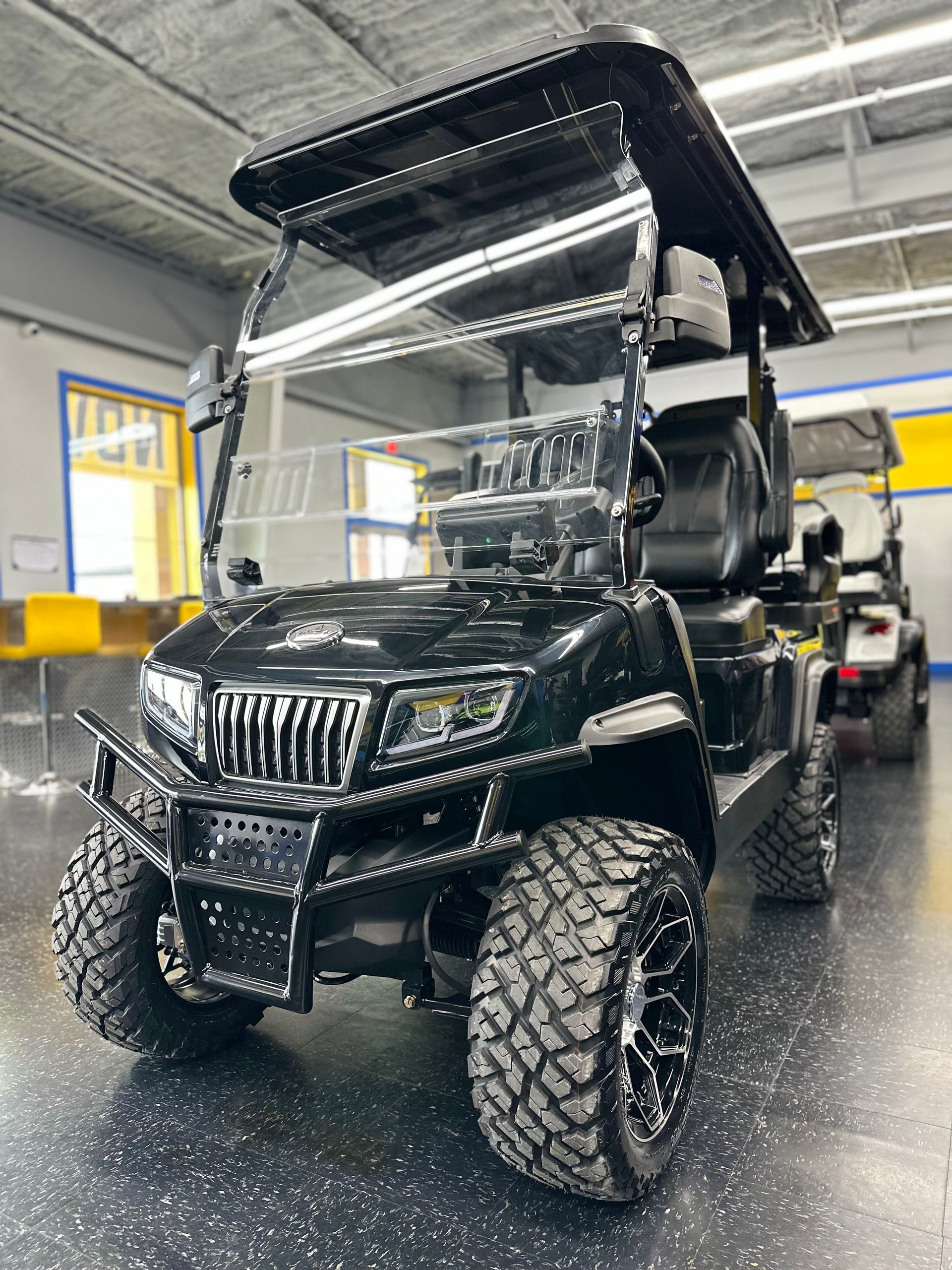 Black golf cart with large off-road tires. It has a front brush guard, windshield, and roof, parked indoors.