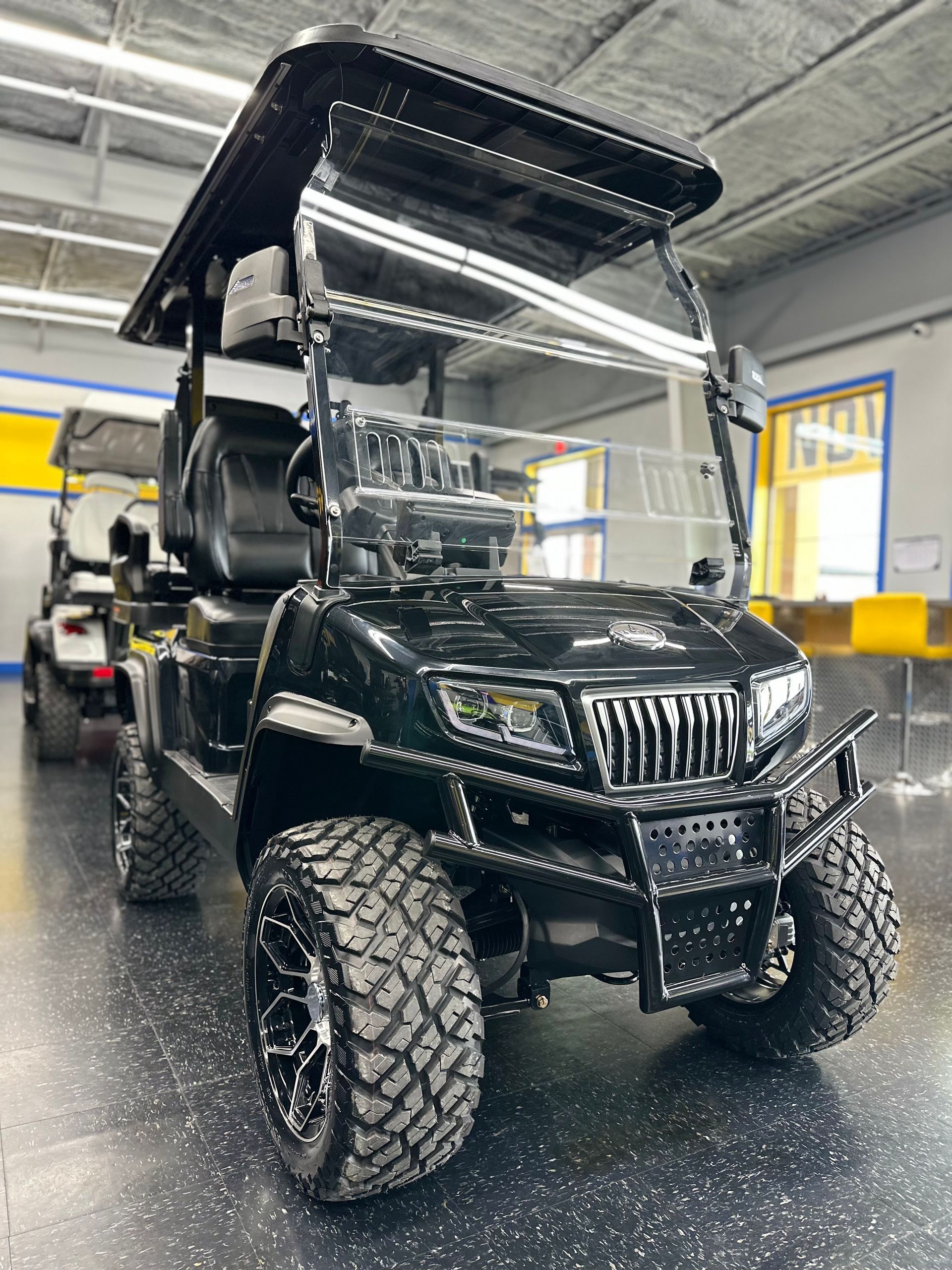 Black golf cart with off-road tires parked inside a garage. It has a clear windshield, a black roof, and a protective bar around the front.