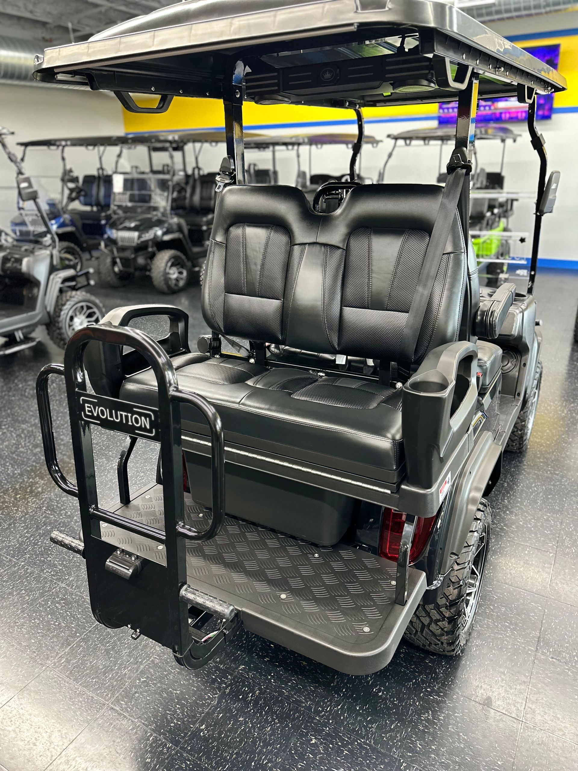 Black golf cart with two rows of seats, rear-facing back seat, and off-road tires, parked indoors in a showroom.
