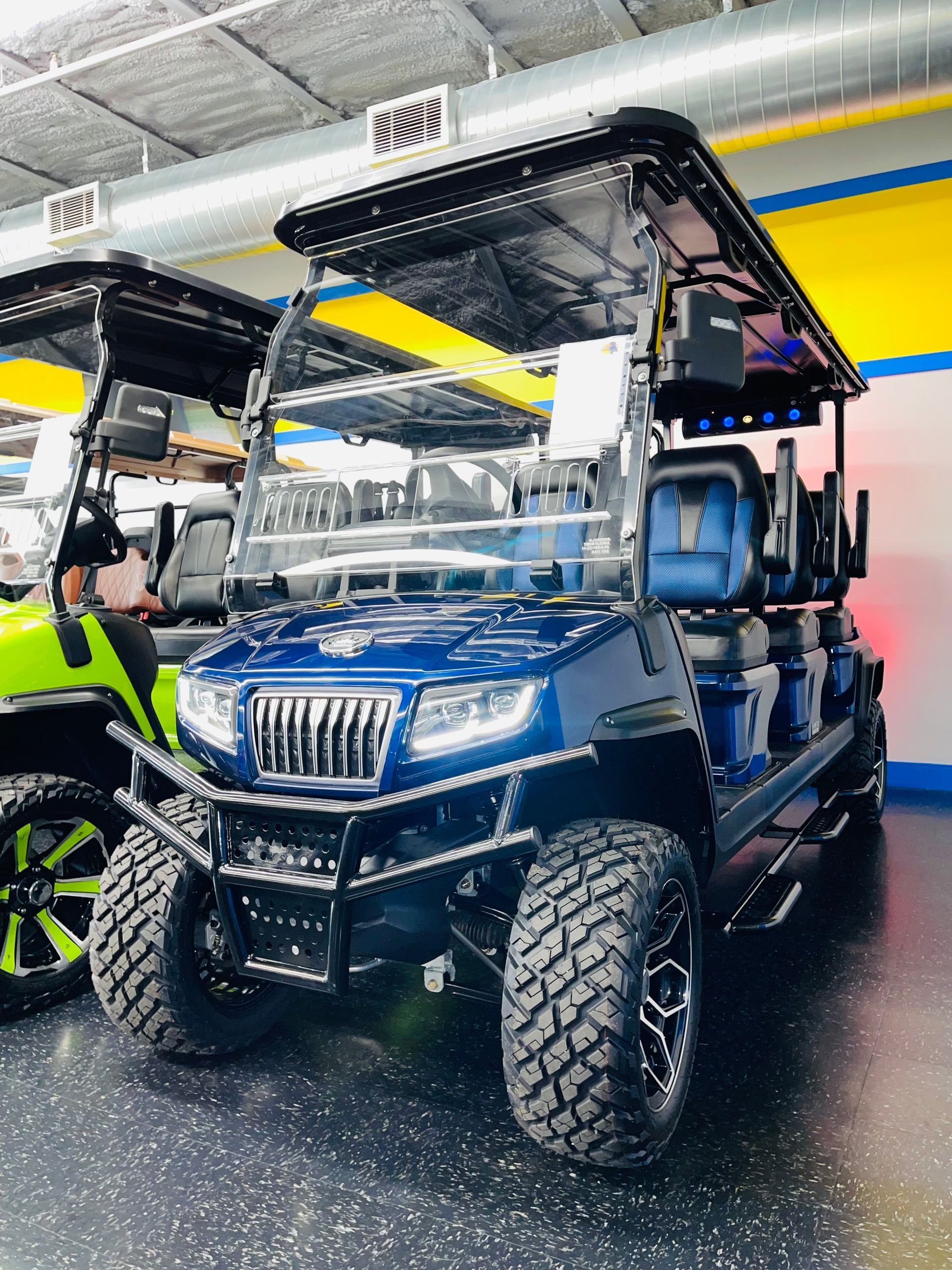 Blue off-road utility vehicle with roof canopy parked indoors, next to a yellow vehicle and a person in neon vest