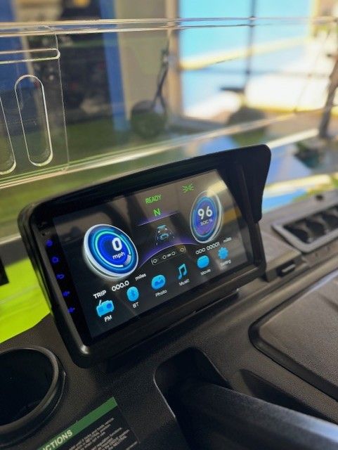 Dashboard of a golf cart displaying speedometer, battery life, and media controls. A black screen with a sun visor is mounted on the cart's interior.