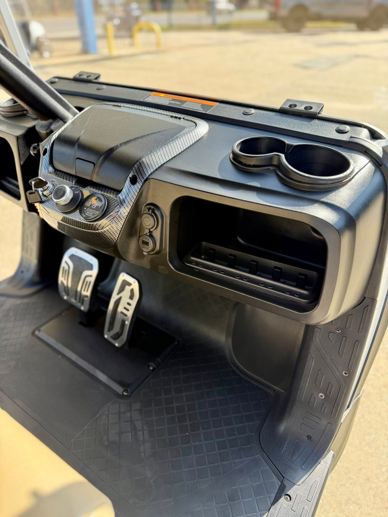 Golf cart dashboard with cup holders, controls, and foot pedals. Black plastic with silver accents.