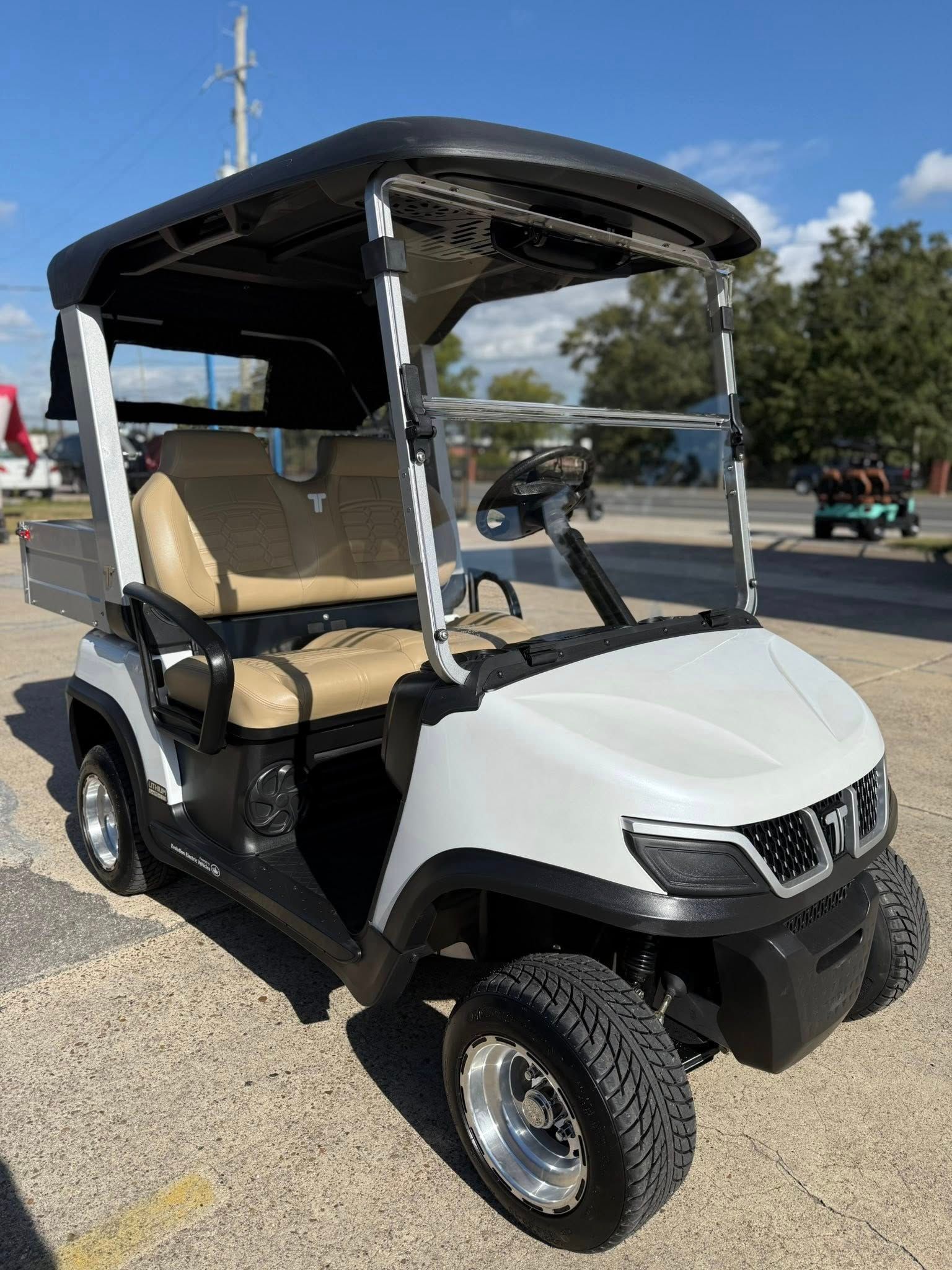White and black golf cart with tan seats parked on pavement, blue sky background.
