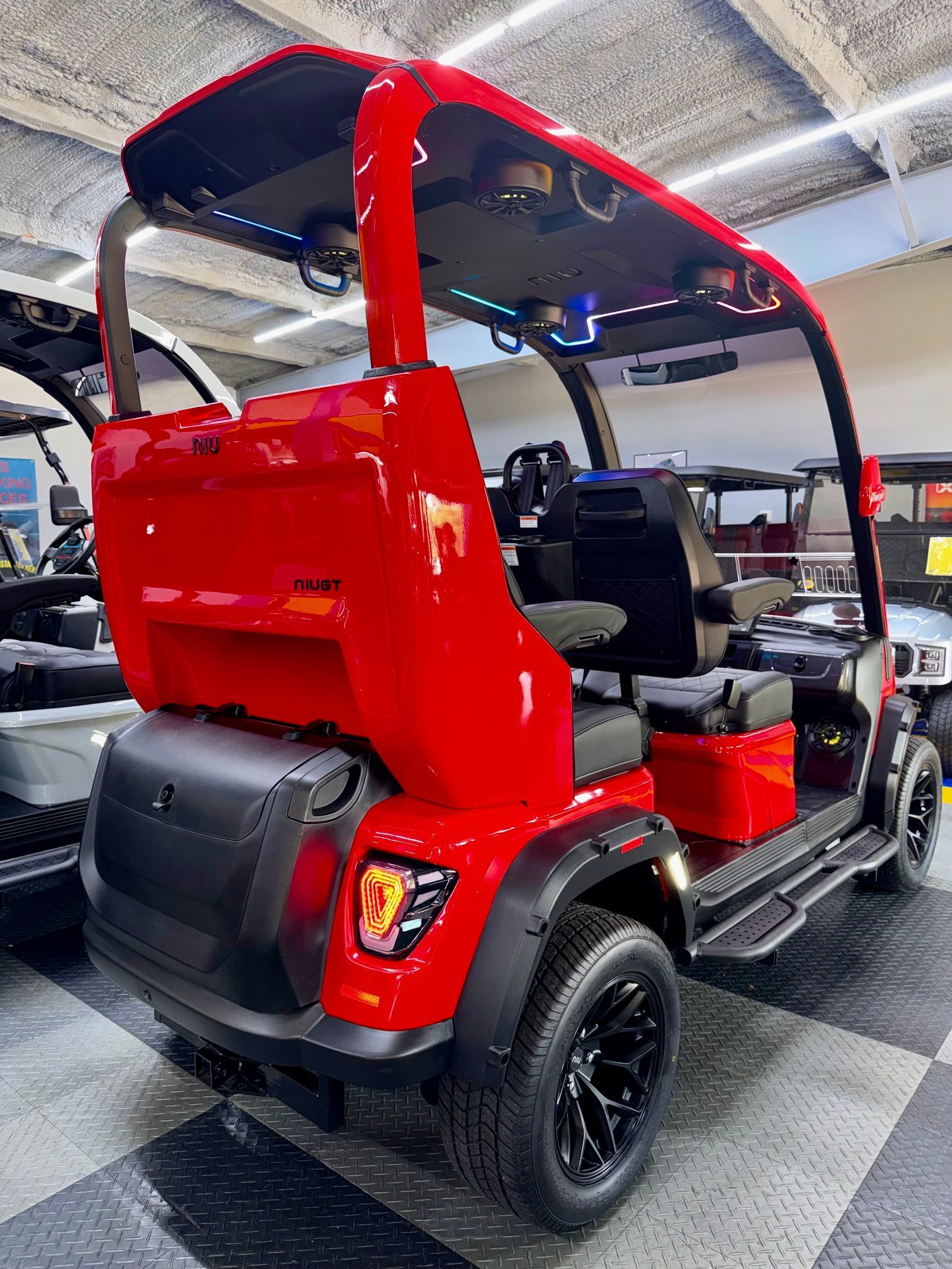 Red electric golf cart with black seats in an indoor showroom, viewed from the rear side.