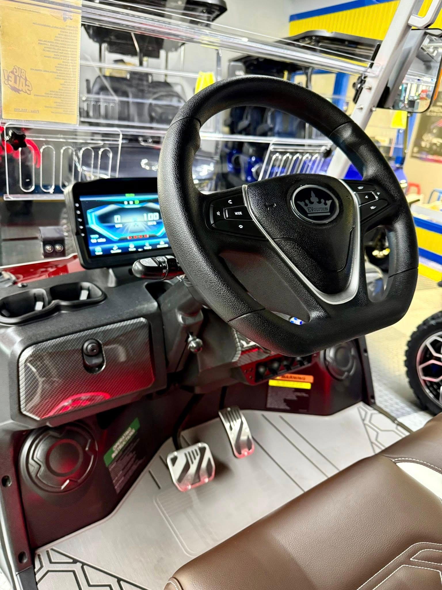 Interior of a custom golf cart with a black steering wheel, digital display, and brown seating. The dashboard features red accents and silver pedals.