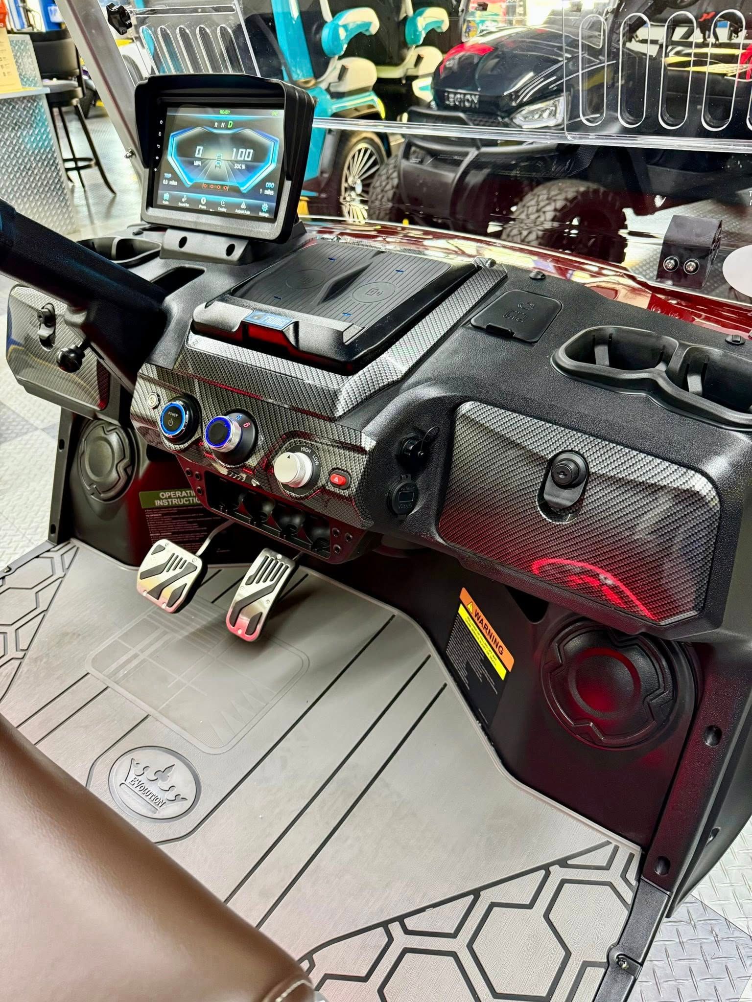 Interior of a customized golf cart. Features a digital dashboard, pedals, speakers, and carbon fiber accents. Brown seat and patterned floor.