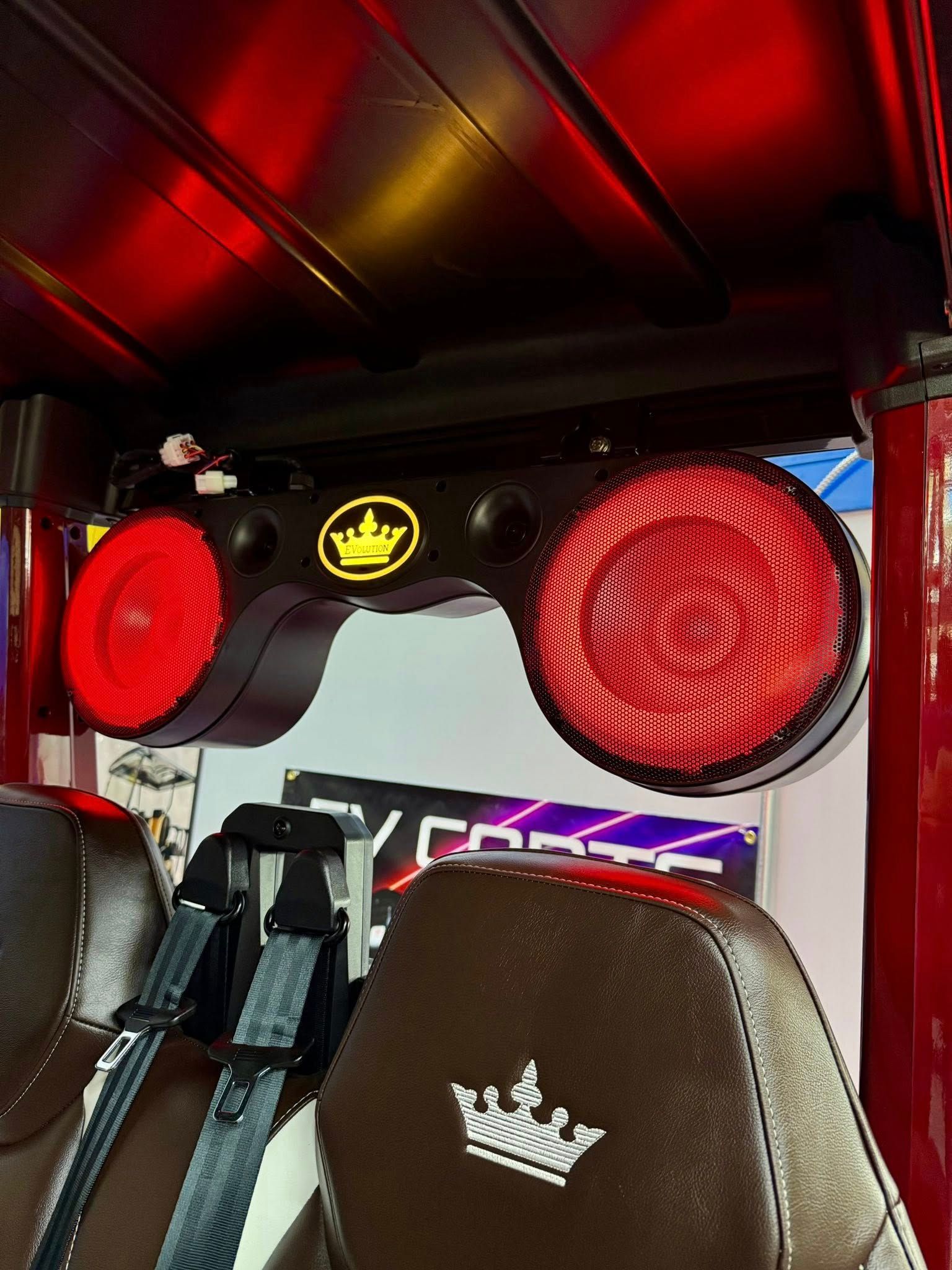 Red-lit speakers mounted in a vehicle's roof. The speaker system has a crown logo, and brown seats are visible.