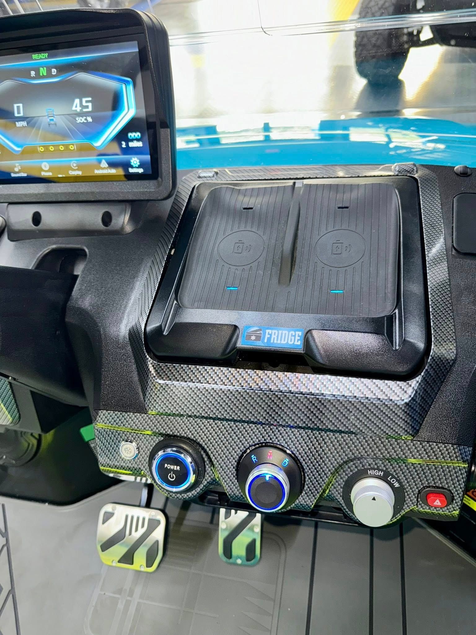 Dashboard of a vehicle with digital displays, climate control knobs, and racing pedals, featuring carbon fiber accents. The speedometer reads 45 mph.