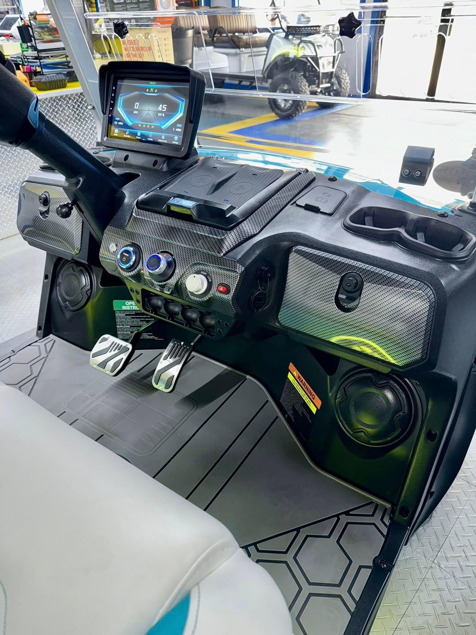 Interior view of a customized golf cart with a digital display, speakers, carbon fiber trim, and metallic foot pedals.
