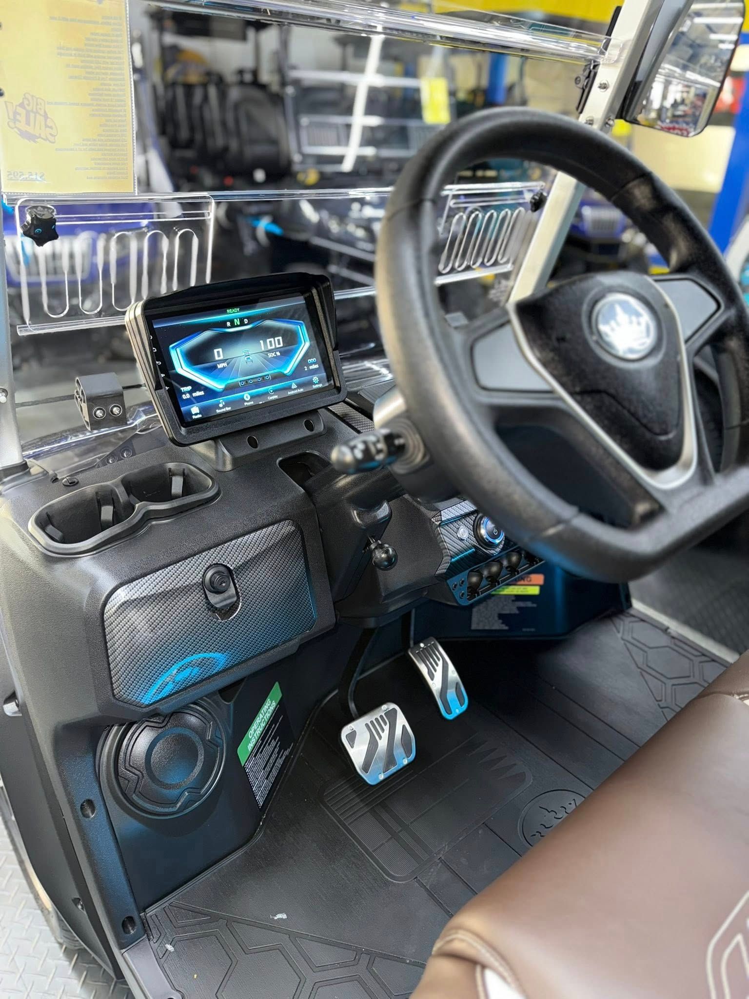 Interior of a golf cart: Black, with steering wheel, touchscreen, pedals, speakers, and cupholders.