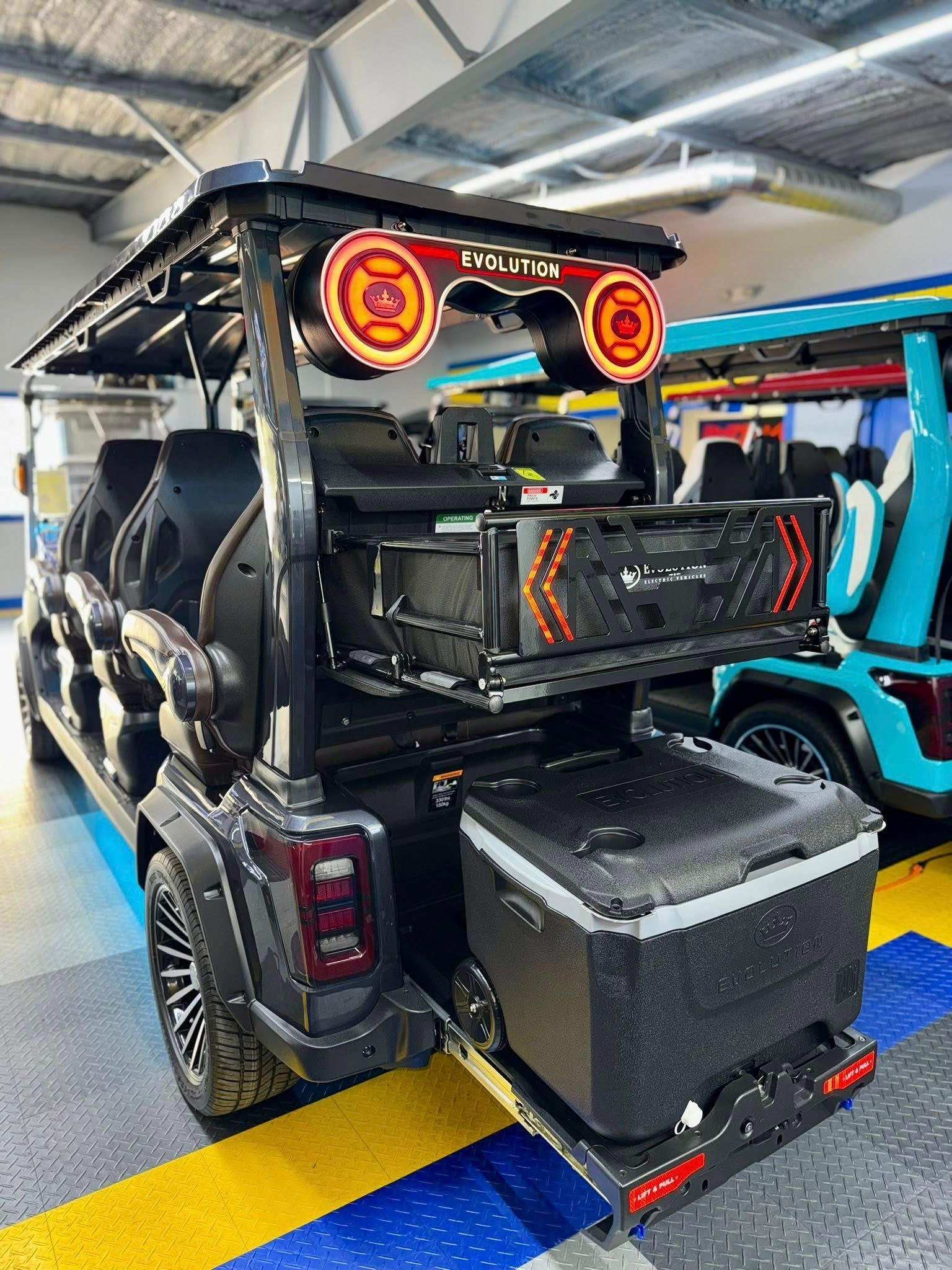 Black, customized golf cart with speaker lights and a cooler, parked indoors next to a blue cart.
