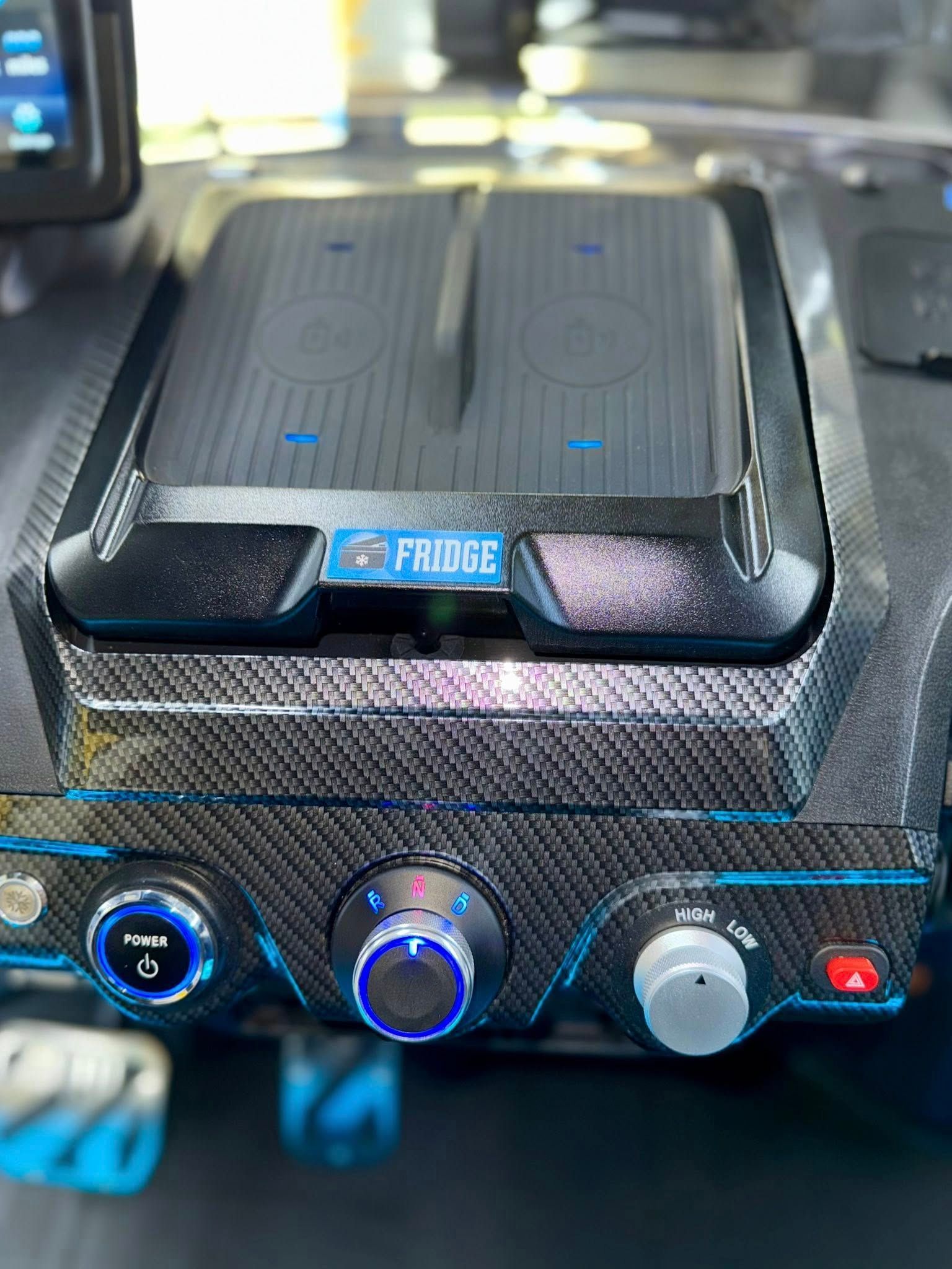 Close-up of a car's console with a carbon fiber look. Features include a fridge, wireless charging, and illuminated controls.