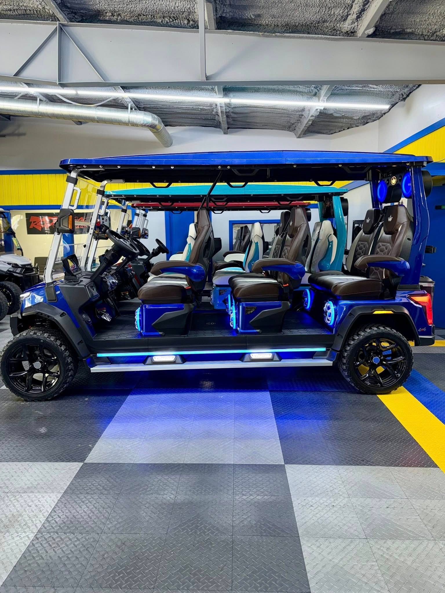 Blue golf cart with black wheels, seating, and roof, parked indoors on checkered flooring.