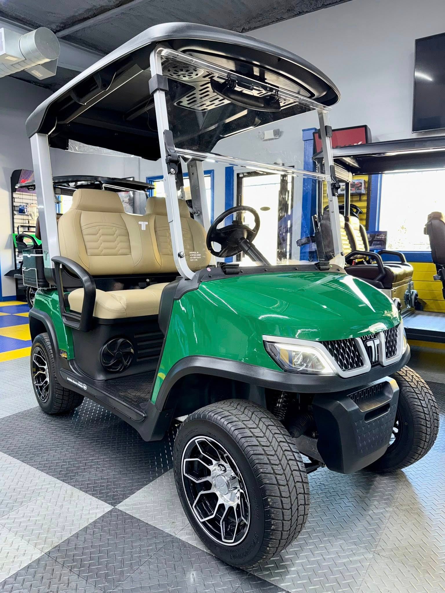 Green golf cart with black accents, tan seats, and off-road tires, inside a showroom.