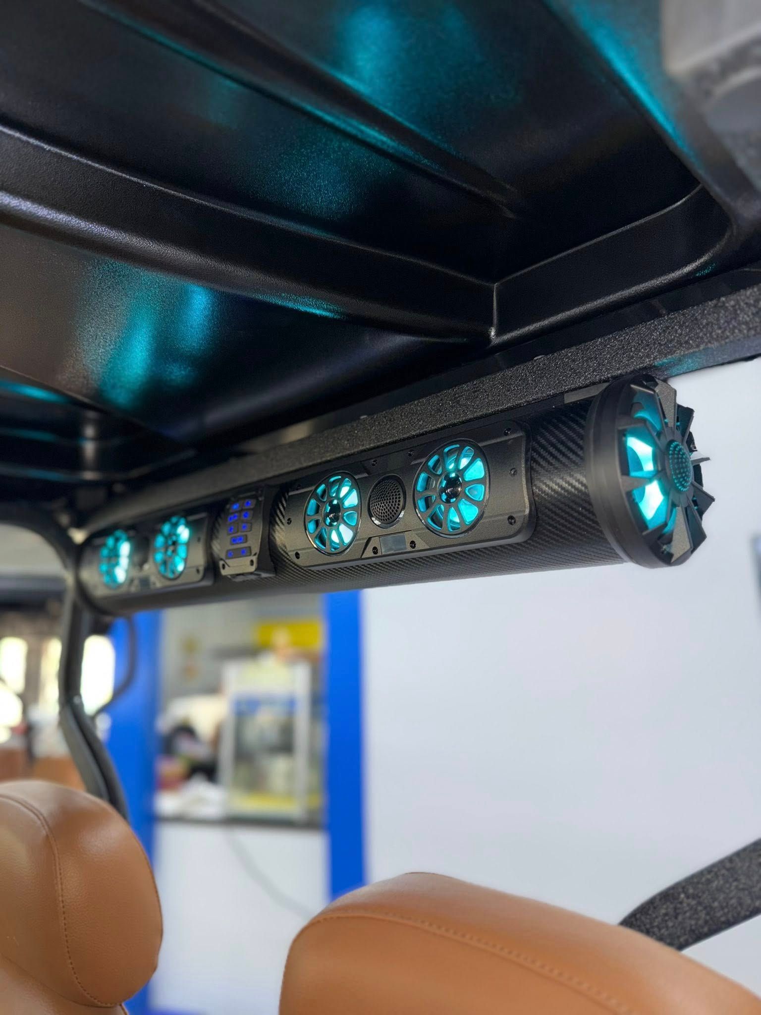 A golf cart's ceiling-mounted speaker system with blue LED lights, seen from inside with tan seats.