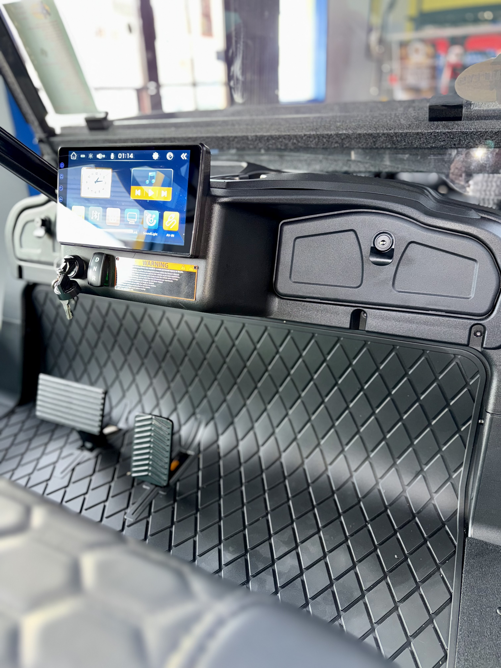Interior of a vehicle with a mounted touchscreen display, pedals, and diamond-patterned mat.