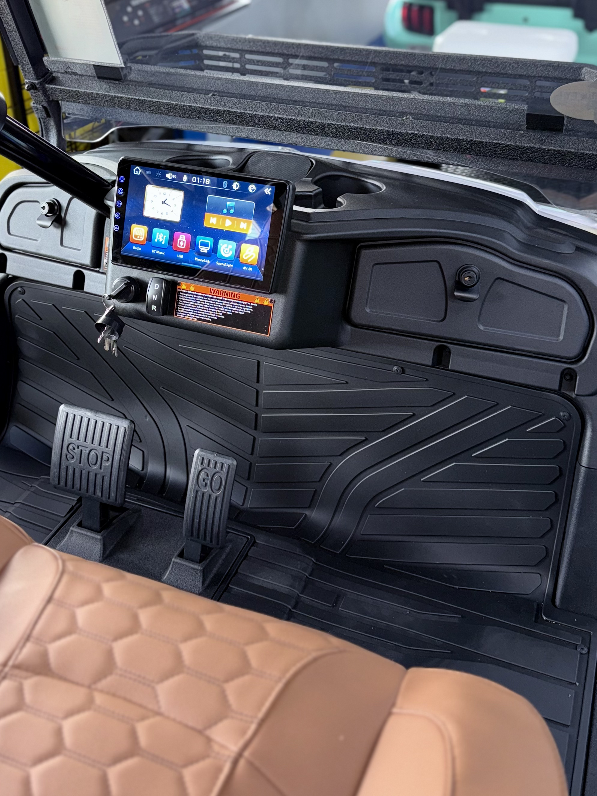 Interior view of a vehicle dashboard with an infotainment screen, featuring brown and black accents, pedals, and a floor mat.