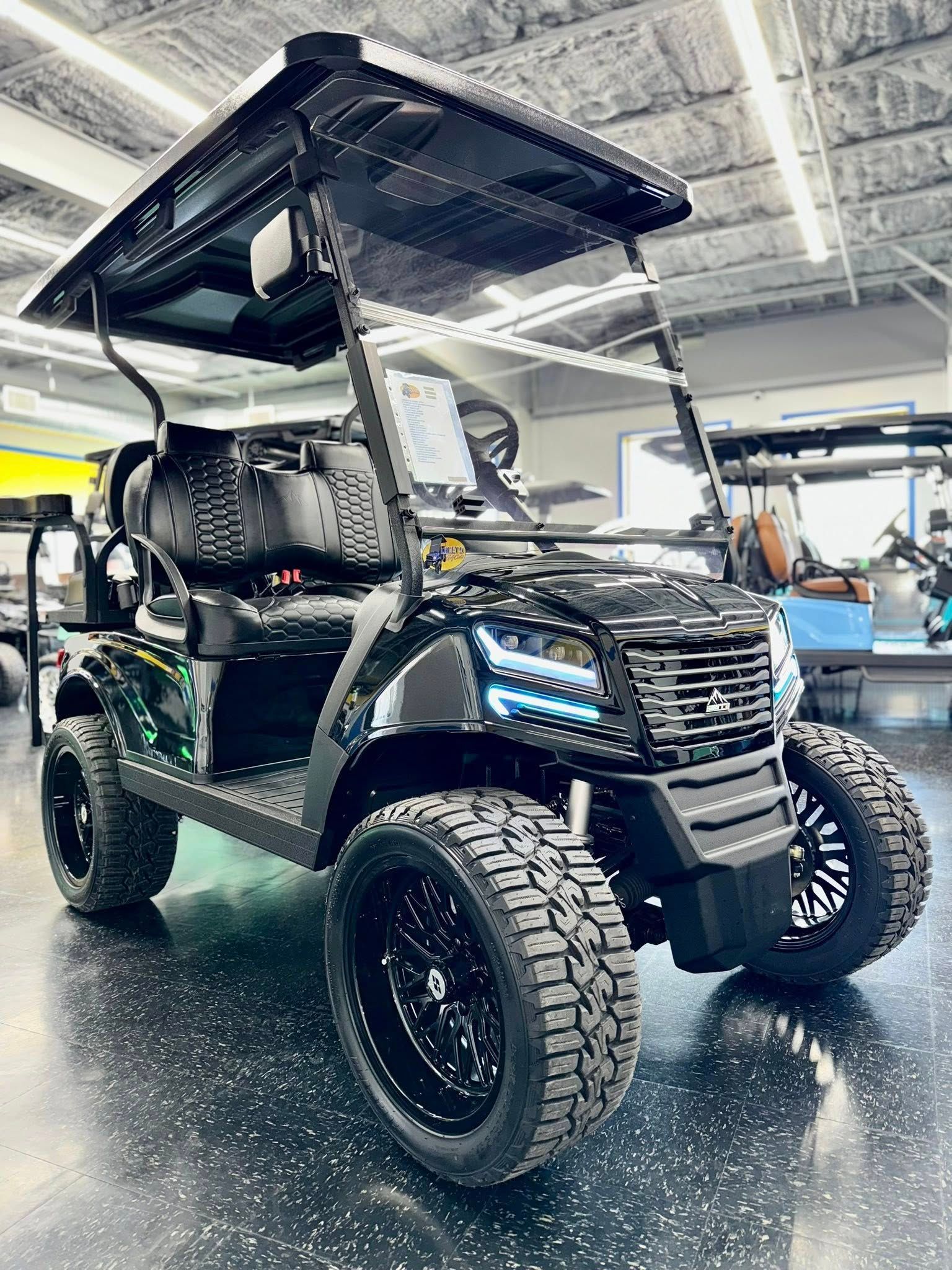 Black lifted golf cart with large tires, glossy black rims, and LED headlights, parked indoors.