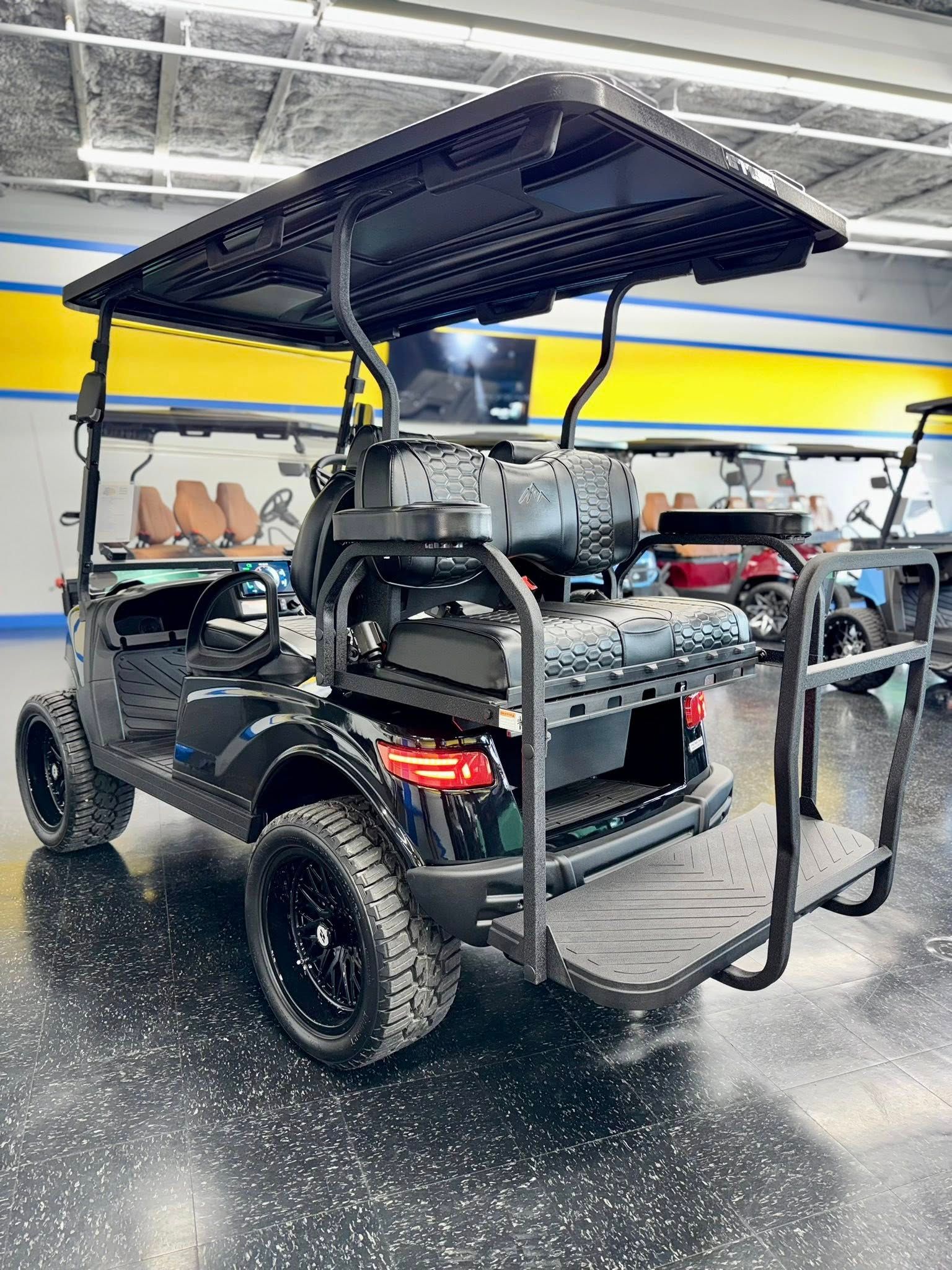 Black golf cart with upgraded wheels, and a rear seat facing backward, indoors.