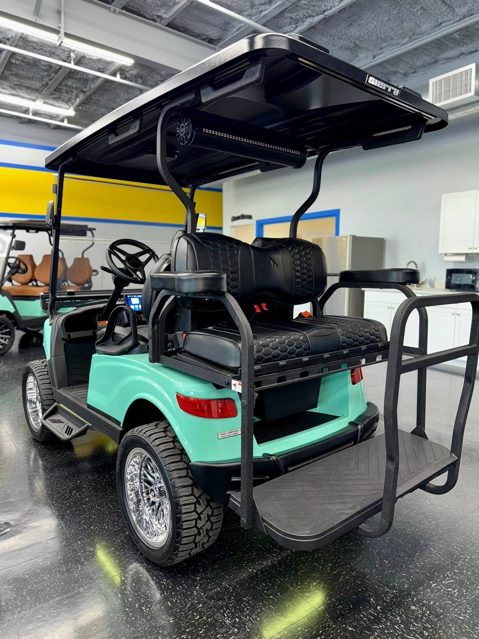 Teal golf cart with black roof, tires, and back platform. Inside a showroom.