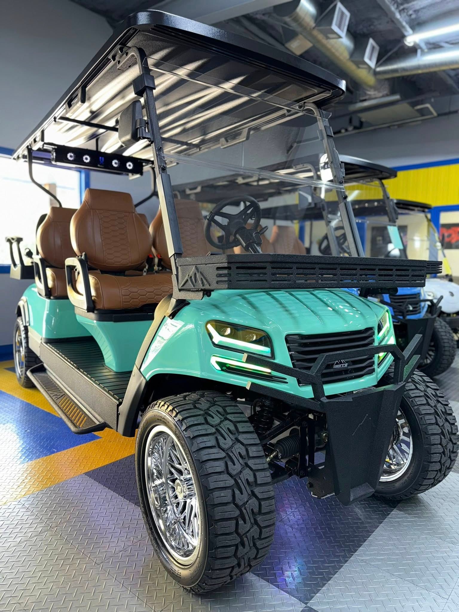 Teal golf cart with brown seats, black trim, chrome wheels, and large tires. Indoors, showroom setting.