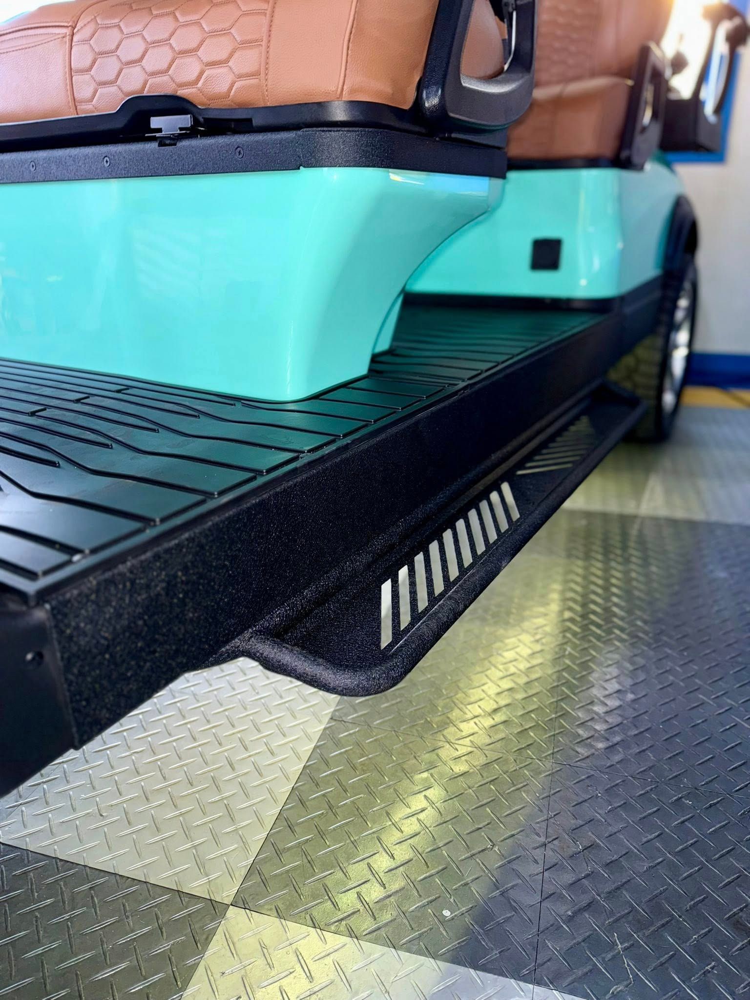 Close-up of a black golf cart side step with silver accent lines. Turquoise and brown seat visible.
