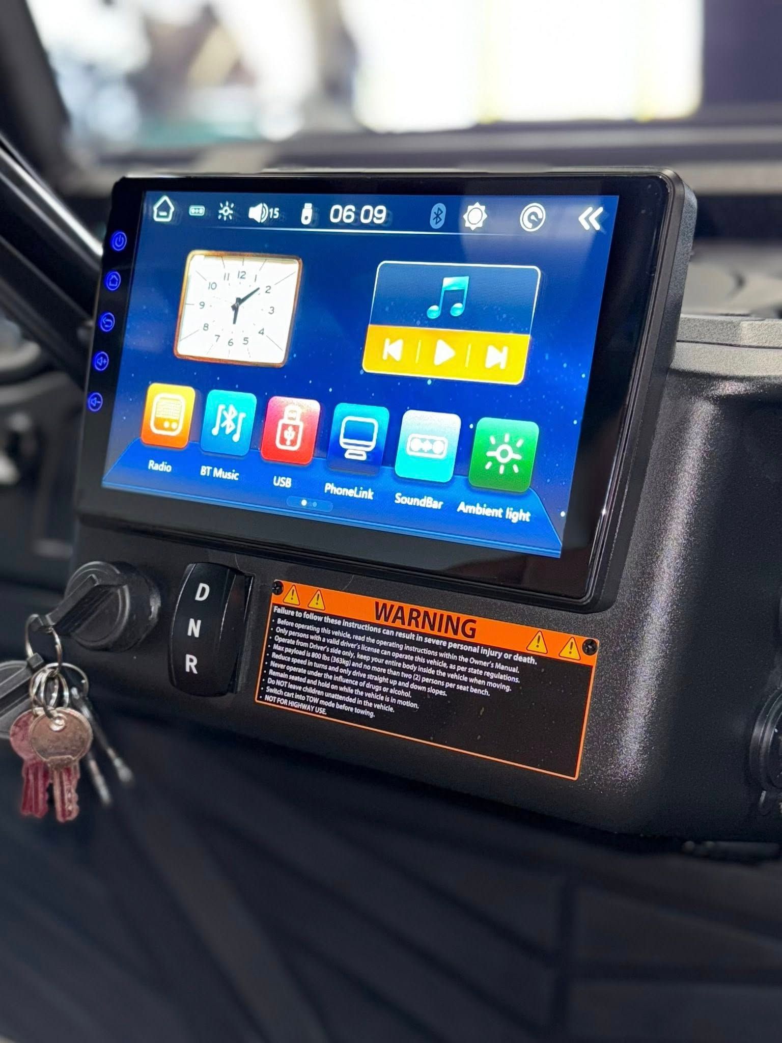 Black car console with touchscreen, showing clock and music app. Keys hanging. Warning label visible.