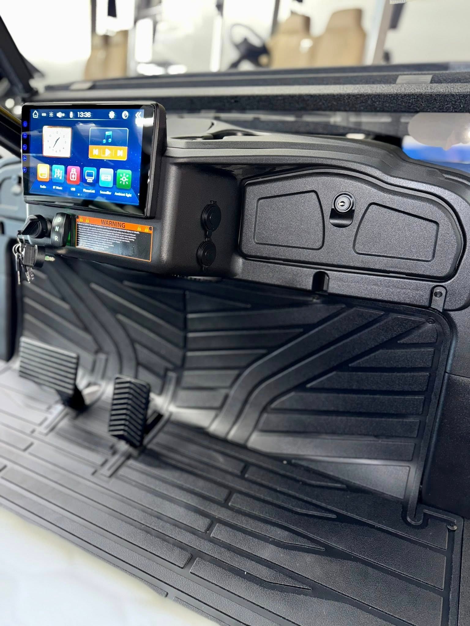 Interior of a vehicle, displaying dashboard with infotainment screen and black floor mats.