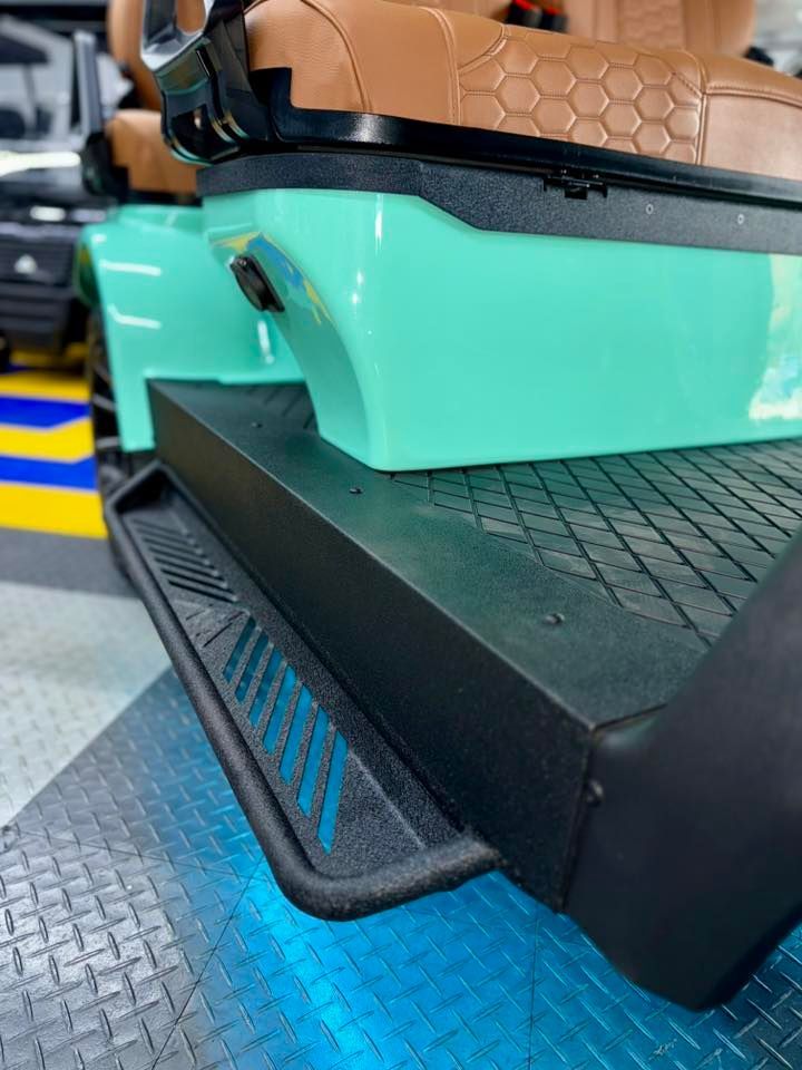 Close-up of a teal golf cart with black steps and a tan seat. The cart is in a garage setting.