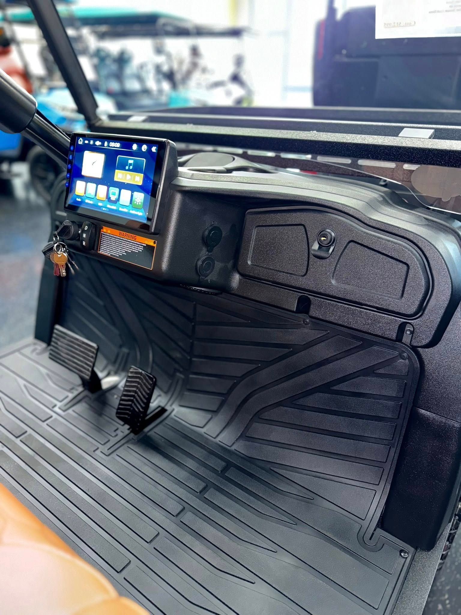 Interior of a vehicle: dashboard with a screen, pedals, and black rubber floor mats.