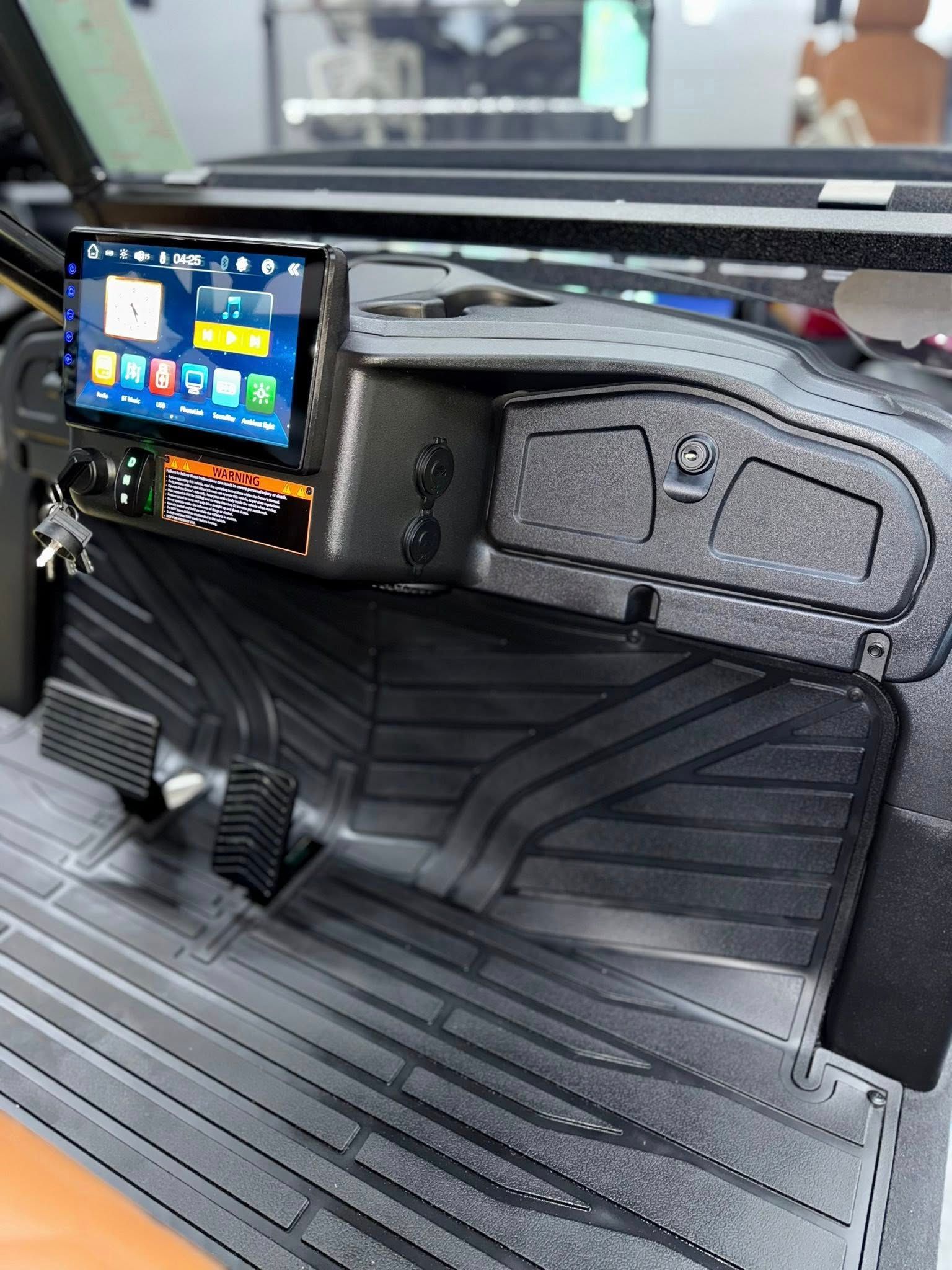 Dashboard of a vehicle featuring a touchscreen, switches, floor mats, and pedals. Black and gray.