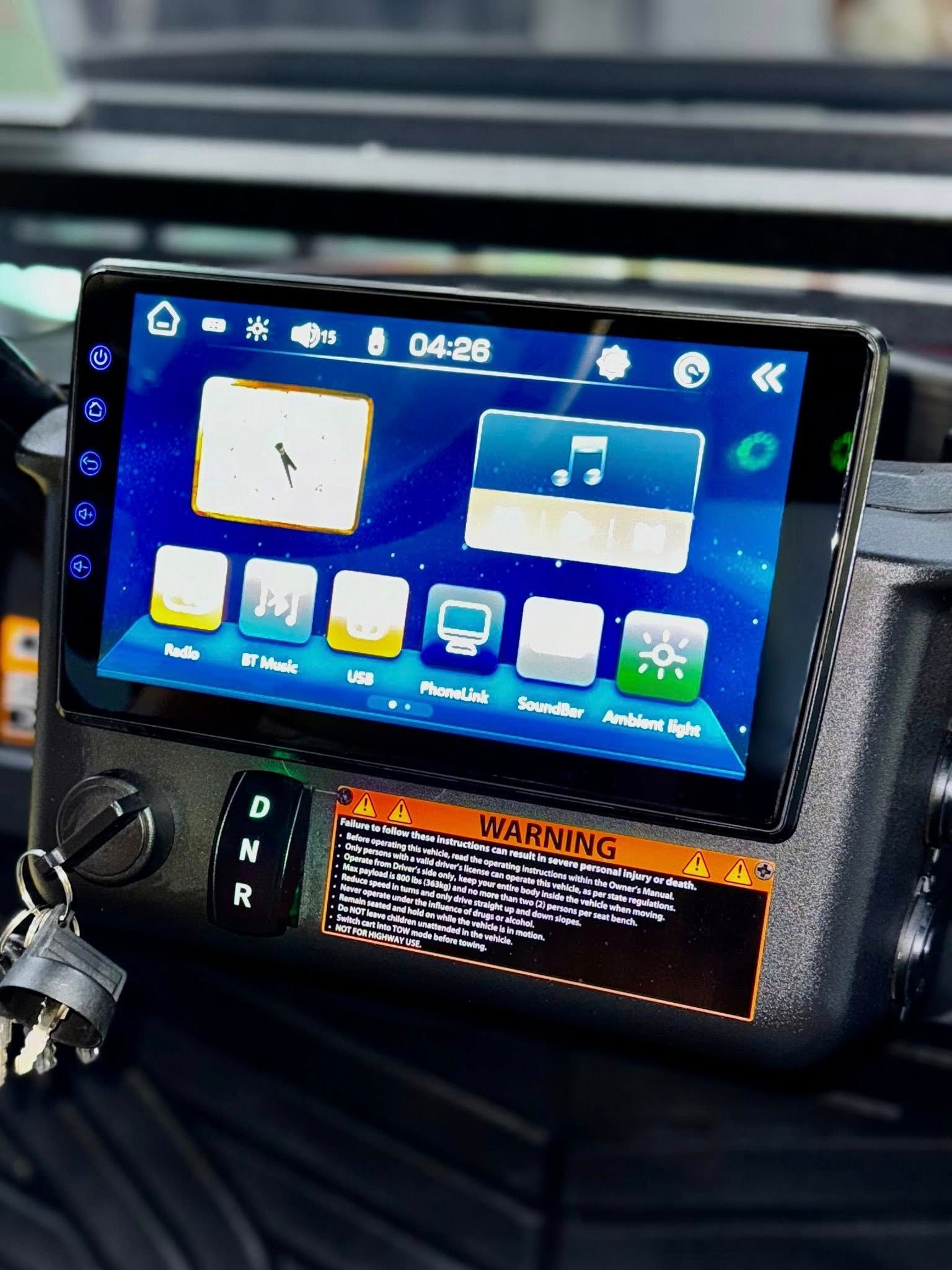 Car dashboard with a touch screen display showing a user interface. Keys are attached. Warning label is visible.