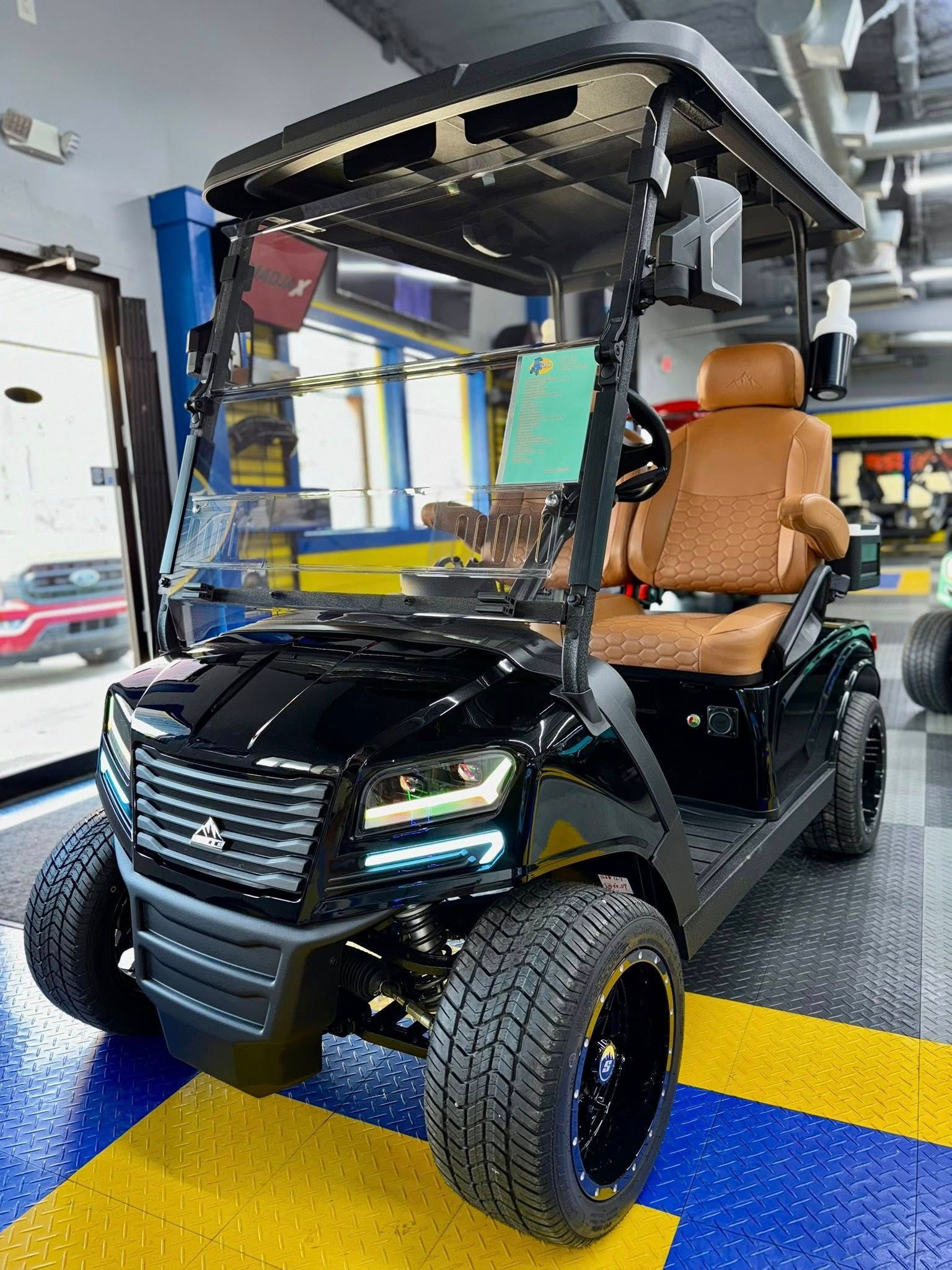 Black golf cart with tan seats, large tires, and a windshield, parked indoors.