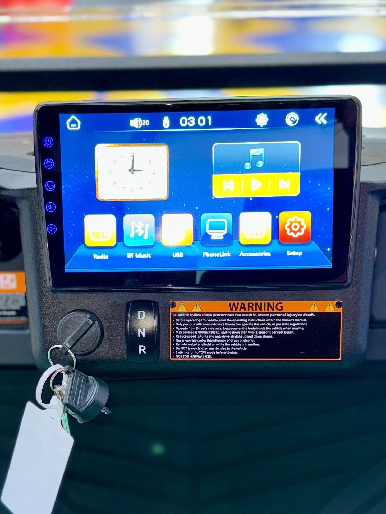 A black car console with a touchscreen display showing clock and music widgets, key in ignition, and warning label.