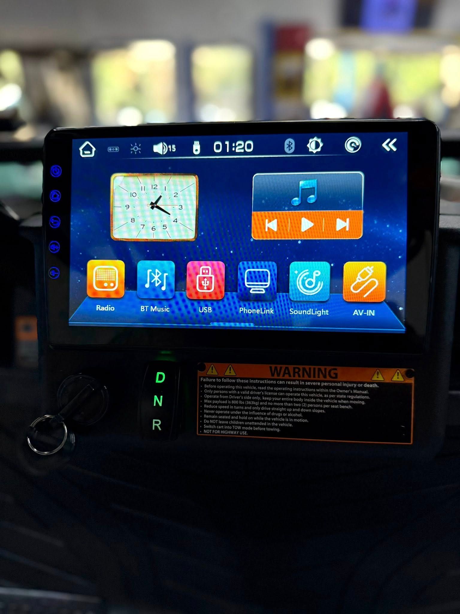 Car dashboard screen showing a clock, music controls, and app icons, with a warning label below.