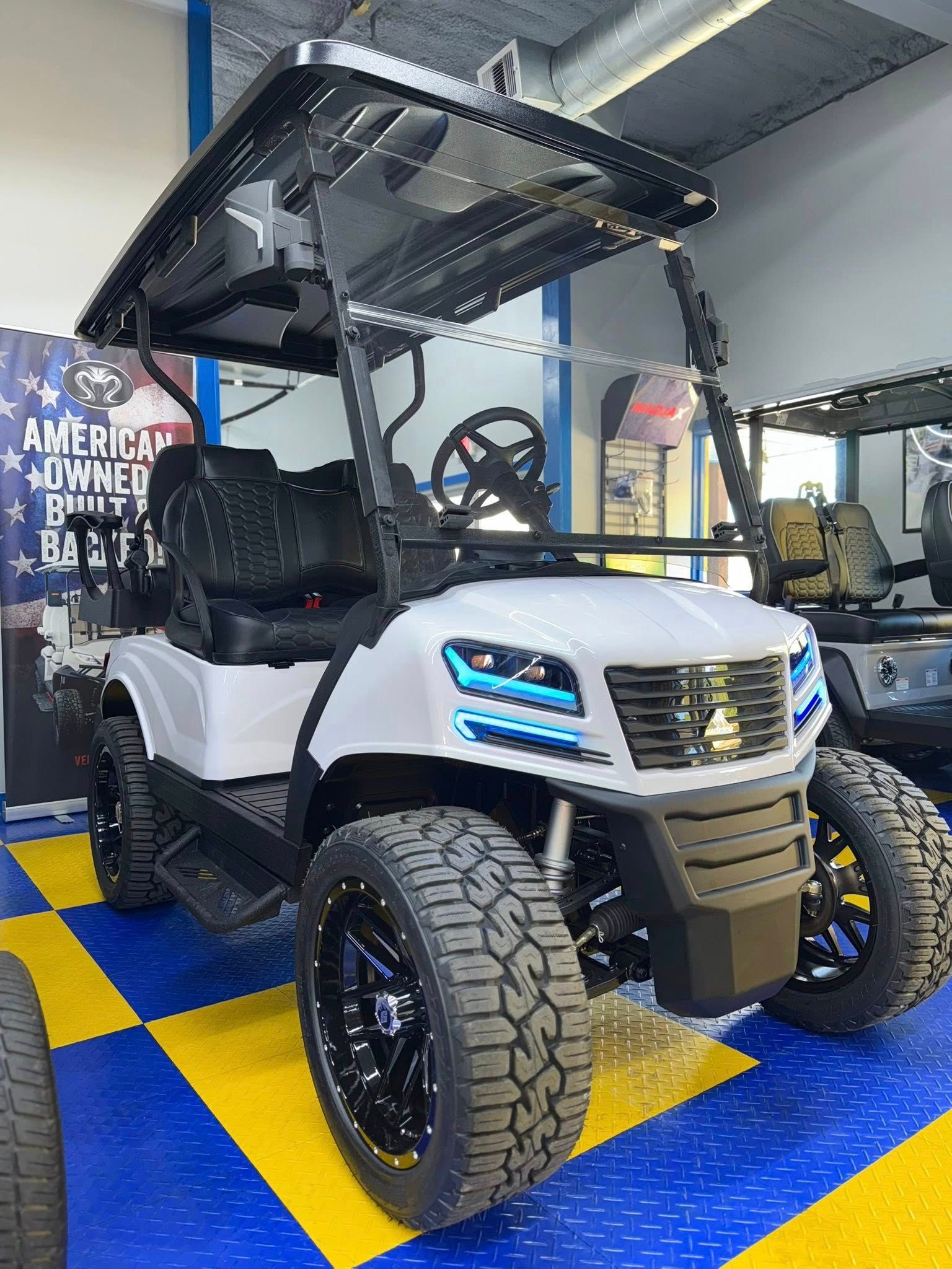 White and black custom golf cart with off-road tires, blue accents, and black roof, parked indoors.