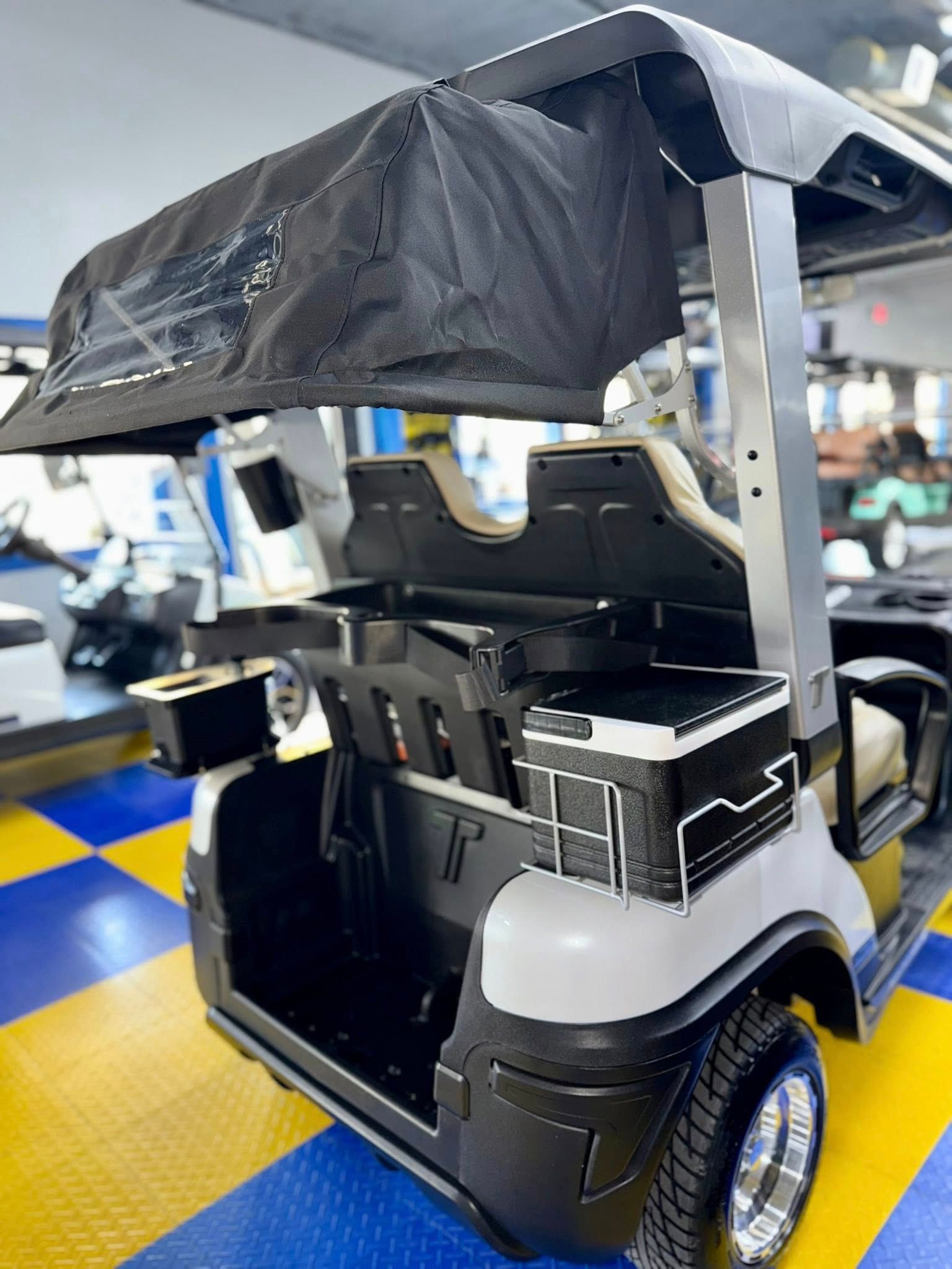 Golf cart with black roof, beige seating, and rear storage compartment, parked indoors.