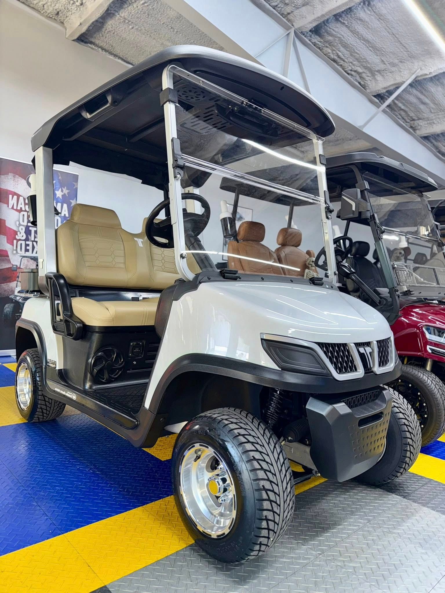 White golf cart with tan seats, black roof, and off-road tires, indoors with another cart.