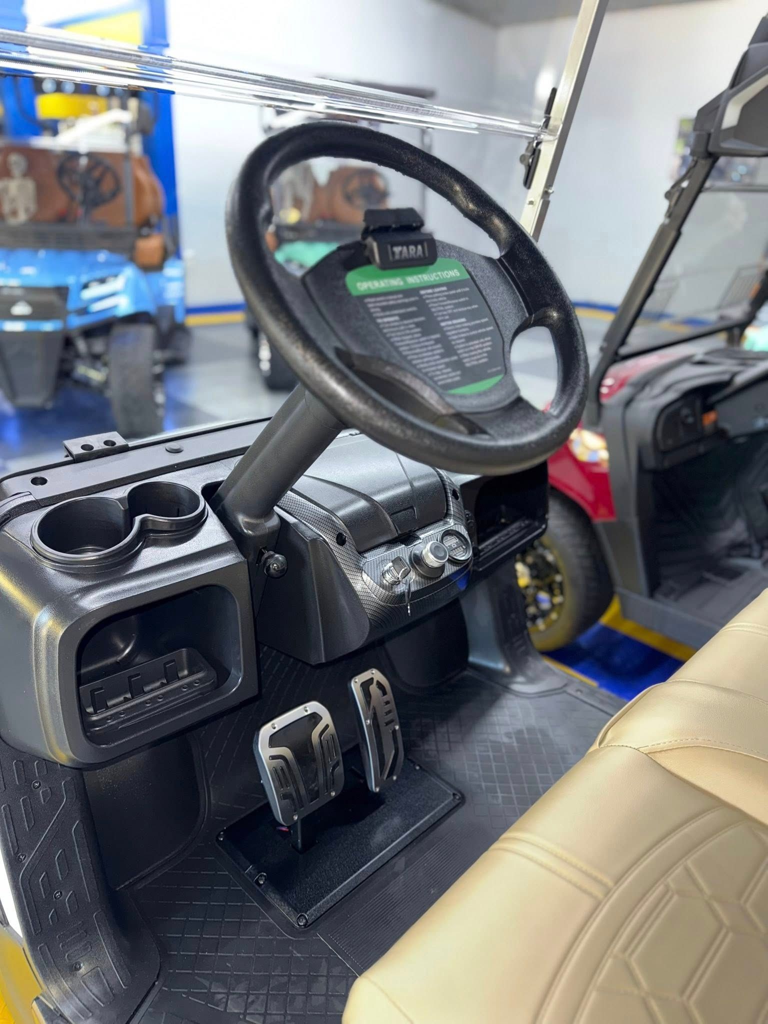 Interior of a golf cart with black dashboard, steering wheel, and tan seat. Another cart in the background.
