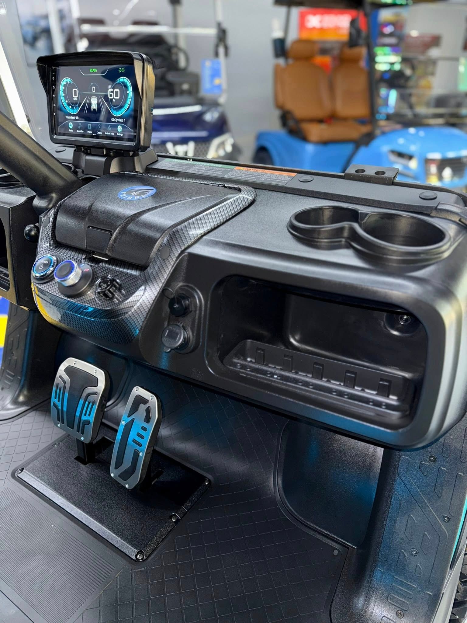 Black golf cart dashboard with cup holders, display screen, pedals, and storage compartment.