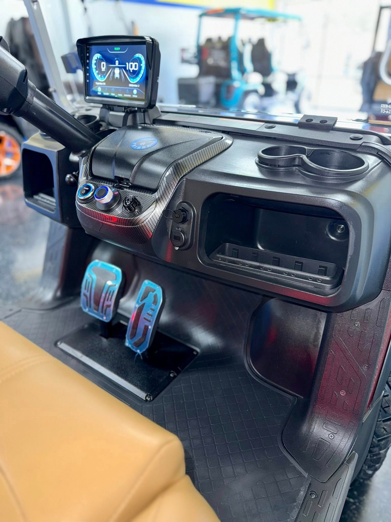 Golf cart interior with digital display, cup holders, and illuminated pedals. Black dashboard with tan seat.