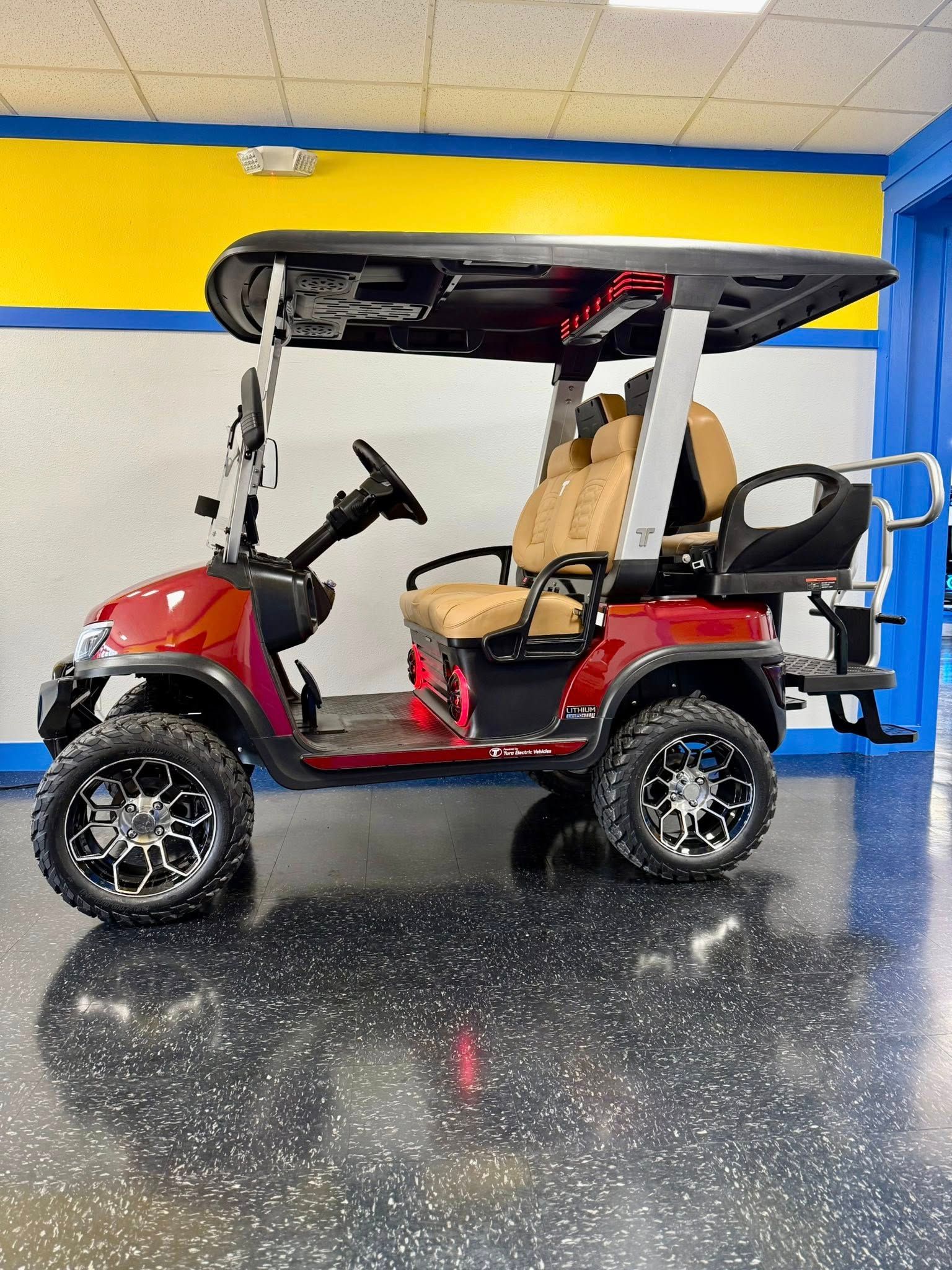 Red and black golf cart with tan seats and black off-road wheels, parked indoors with a yellow wall.